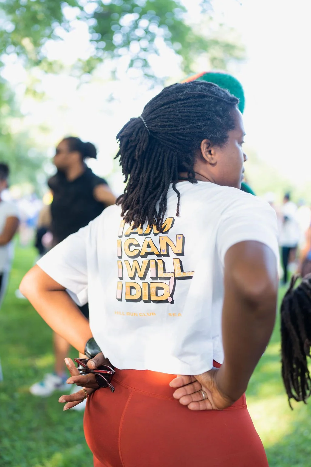 A woman standing outdoors with other people, wearing a white t-shirt with motivational text and red pants, with her hands on her hips, holding sunglasses in one hand, and participating in a group activity or event.