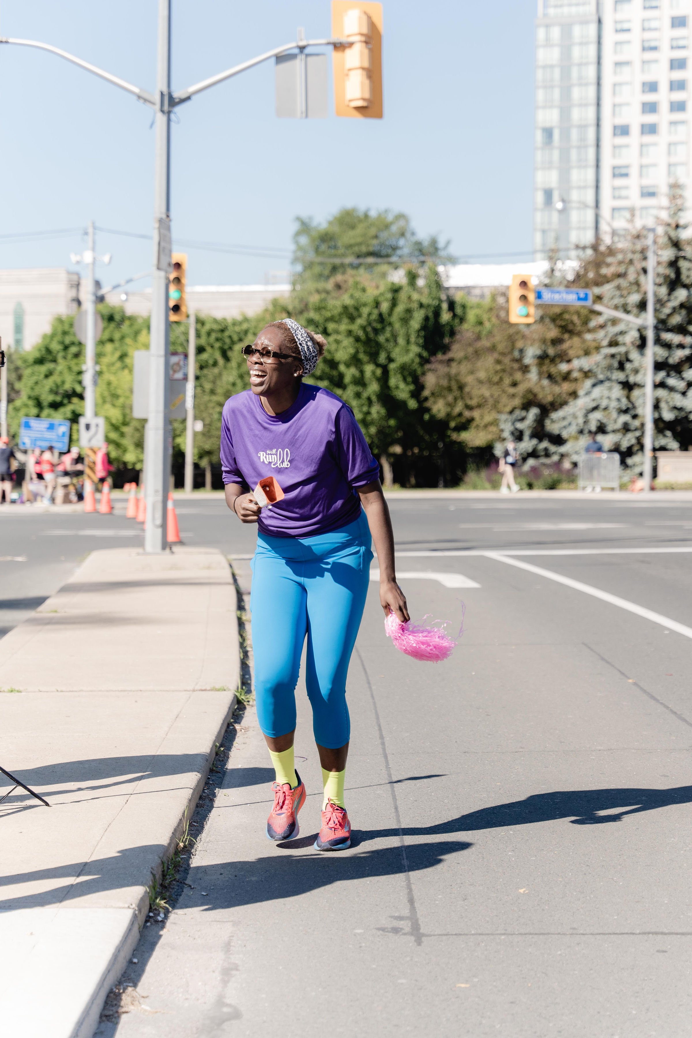 Woman wearing a purple shirt, blue leggings, and pink running shoes participating in an outdoor event, holding a pink feathered accessory and a small orange object, on a city street with traffic lights, trees, and high-rise buildings in the backgroun