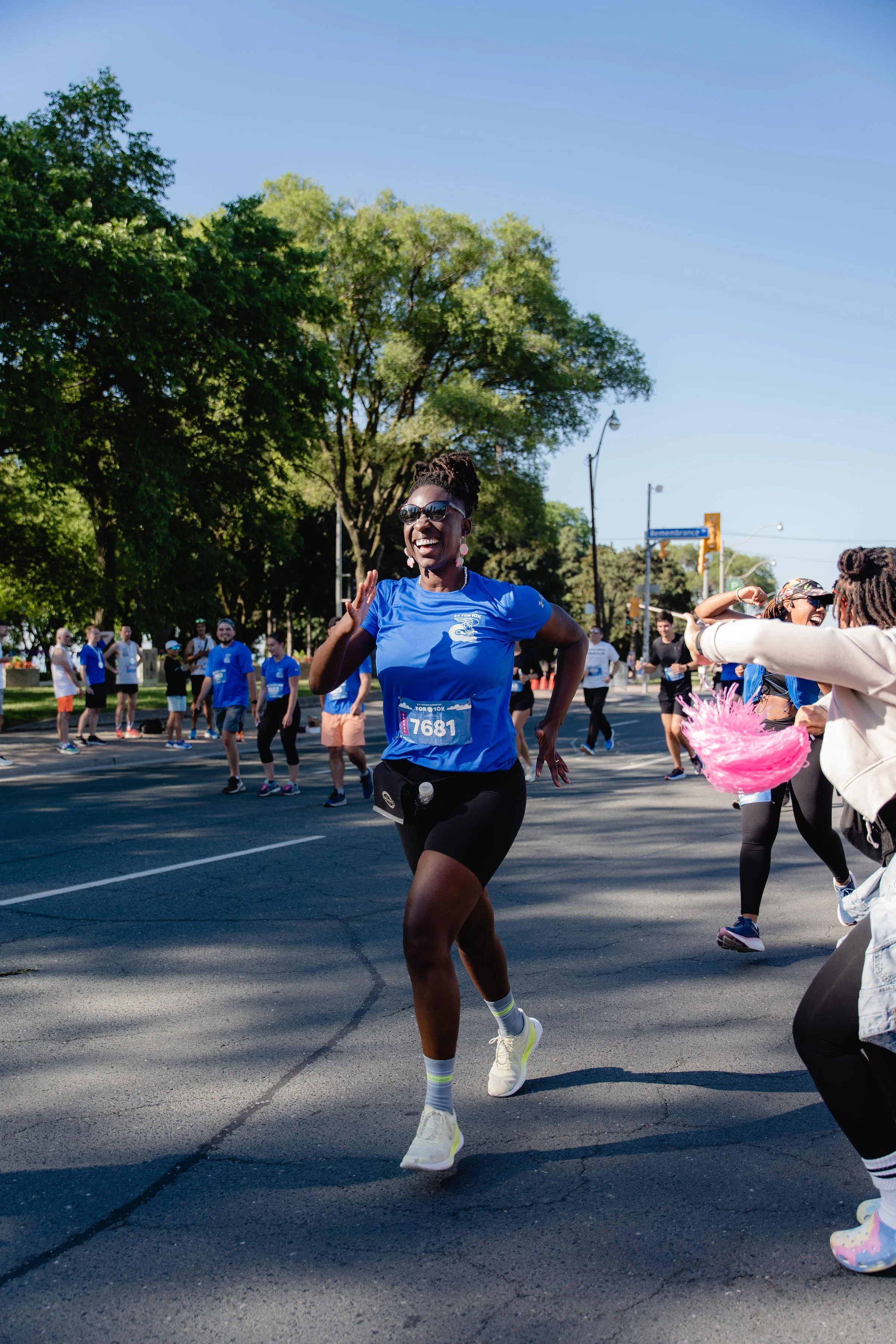 A woman running in a race wearing a blue shirt, black shorts, and sunglasses, with a crowd of people and green trees in the background.