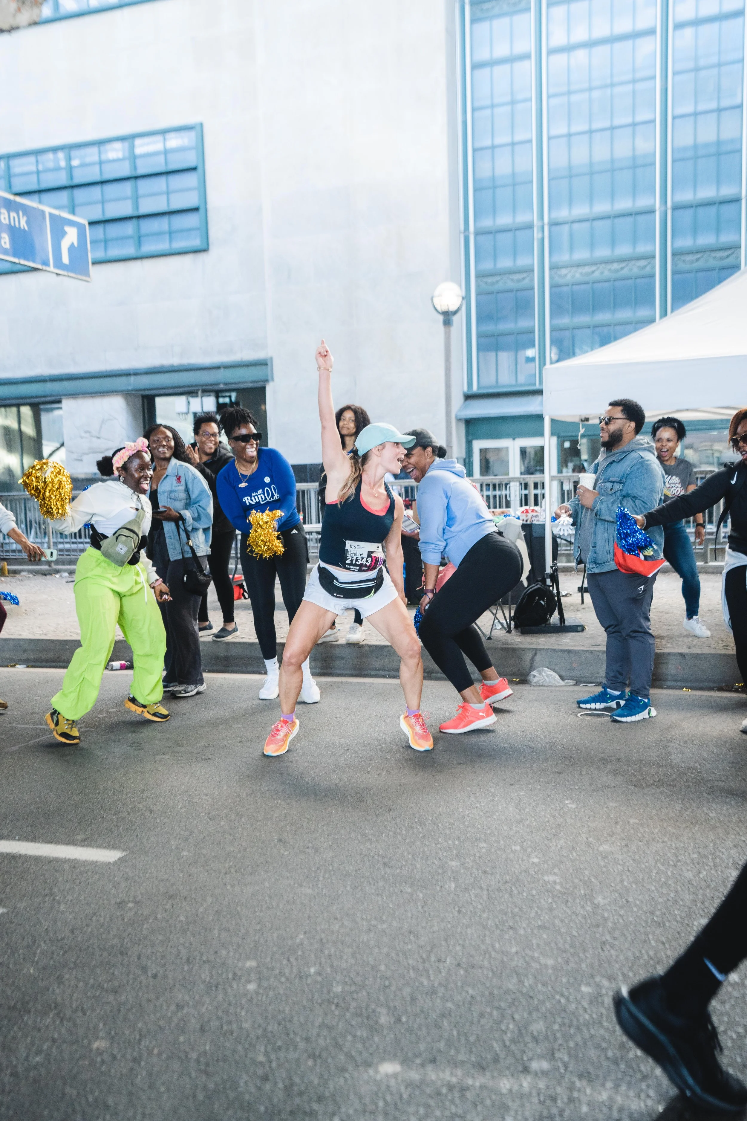 Women dancing and celebrating during a marathon event outside a modern building with a tent and cheerleaders in the background.