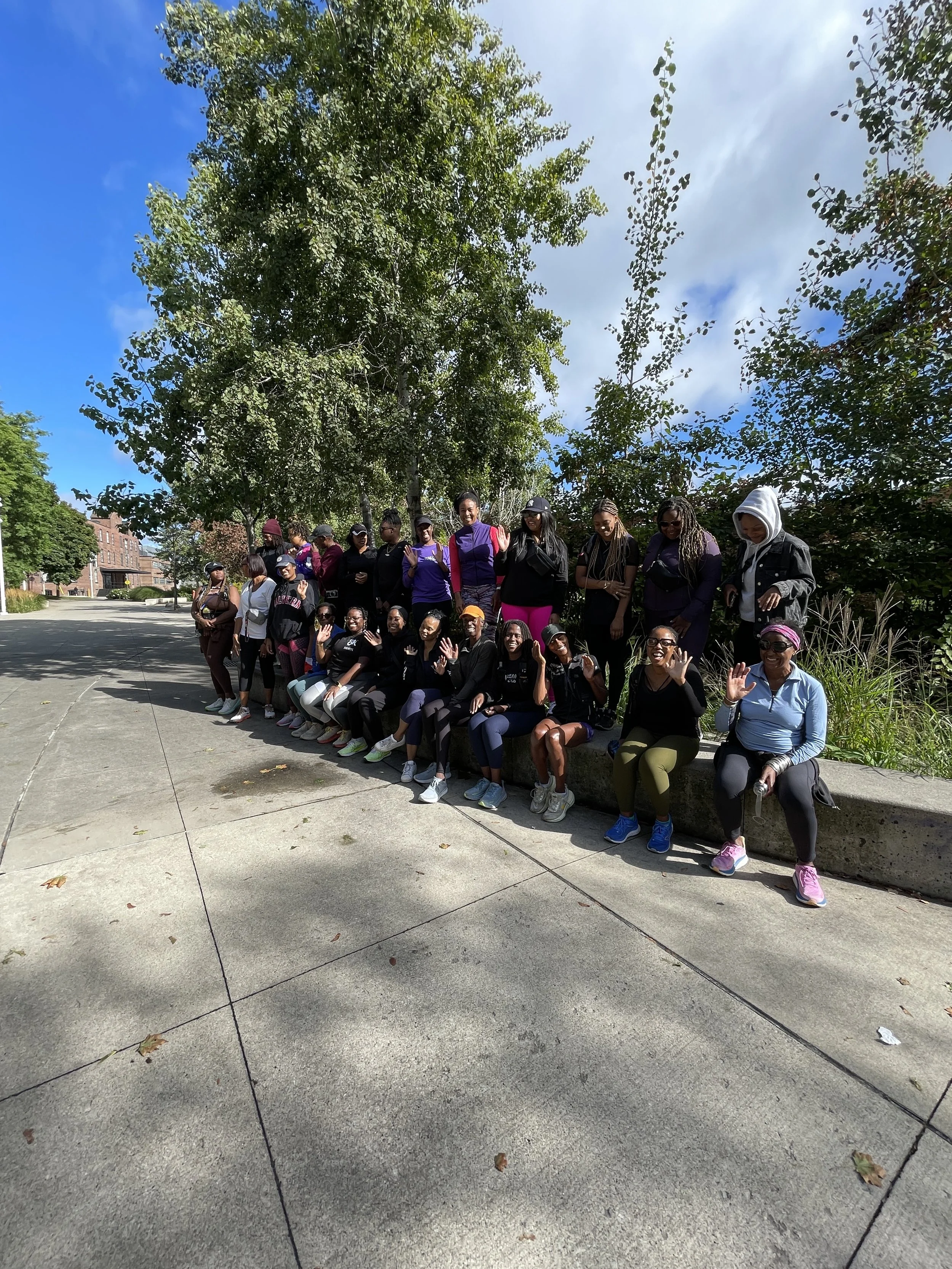 Group of women sitting and standing on a sidewalk near trees, waving at the camera under partly cloudy sky.