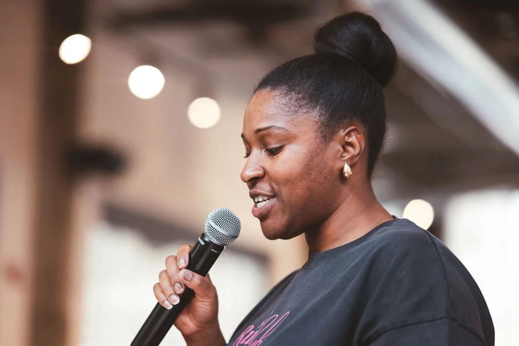 A woman with dark hair tied in a bun speaking into a microphone at an indoor event.