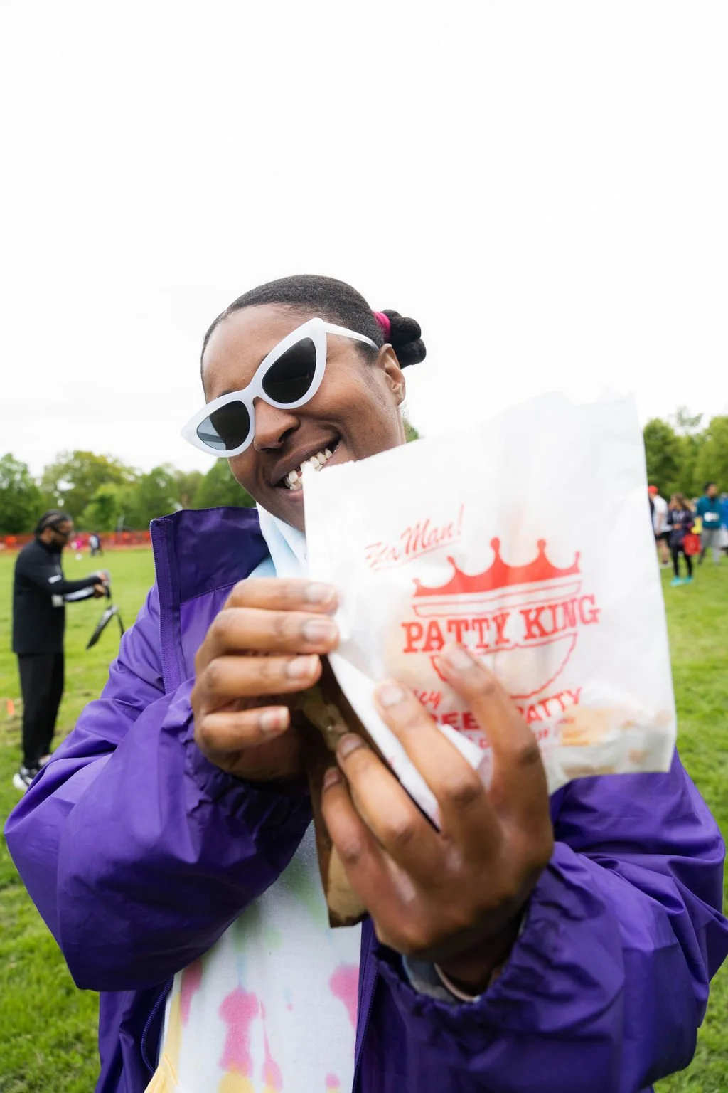 A smiling person wearing white sunglasses and a purple jacket holding a Patty King bag outdoors with other people in the background.