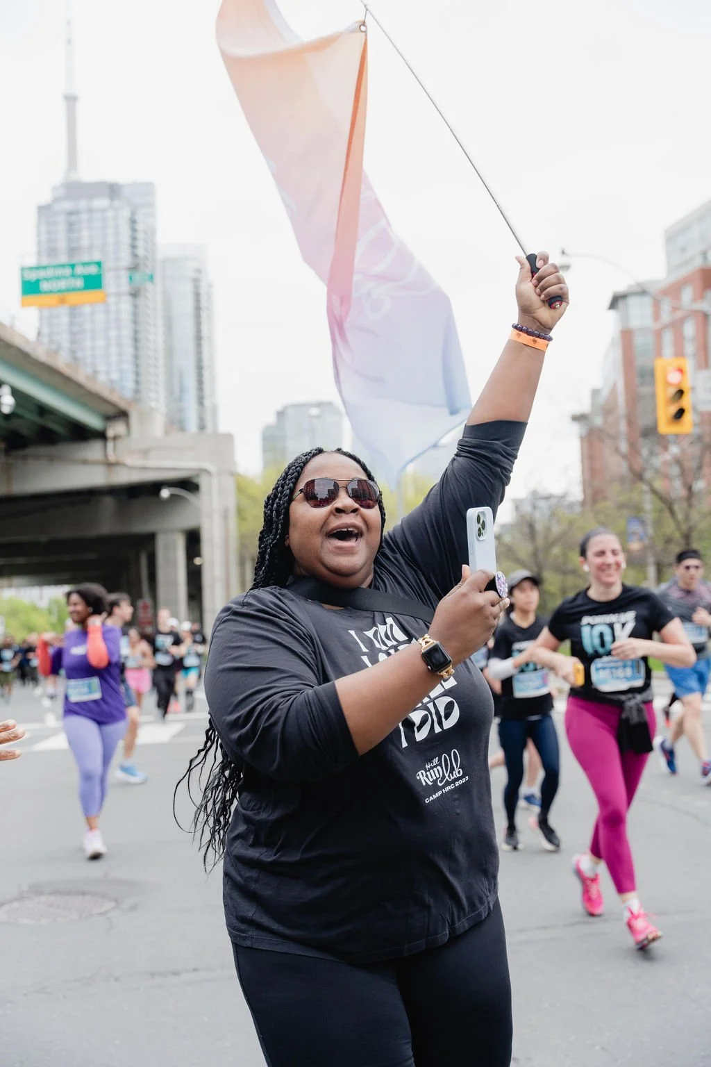 A woman smiling and holding a flag while taking a selfie at a marathon race in an urban area with tall buildings in the background.