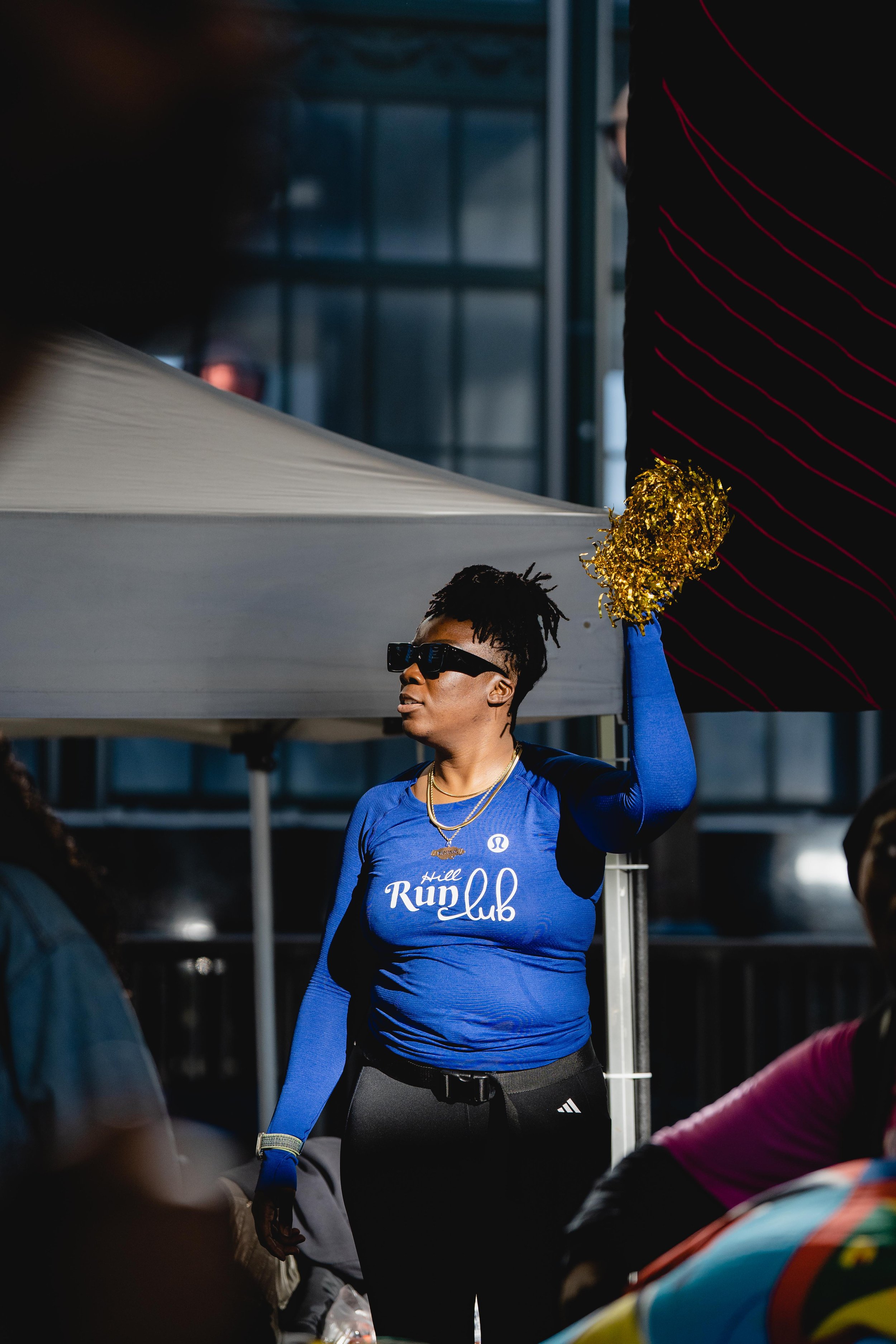 A woman wearing sunglasses and a blue long-sleeve shirt with the words 'Hill Run Club' written on it holds gold pom-poms and is standing outdoors, possibly at a running event or gathering, under a tent.