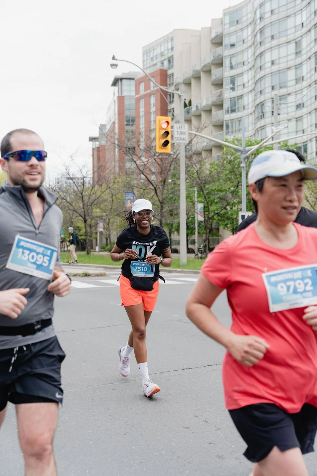 Group of runners participating in a race on a city street, with buildings, trees, and a traffic light in the background.