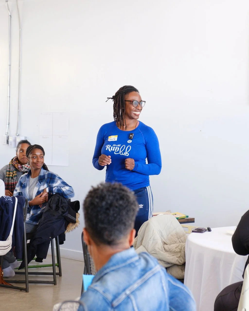 A woman with glasses and dreadlocks smiling, standing in front of a white wall, wearing a blue long-sleeve shirt that says 'Runc Klub' and dark athletic pants, giving a presentation or speech to an audience.