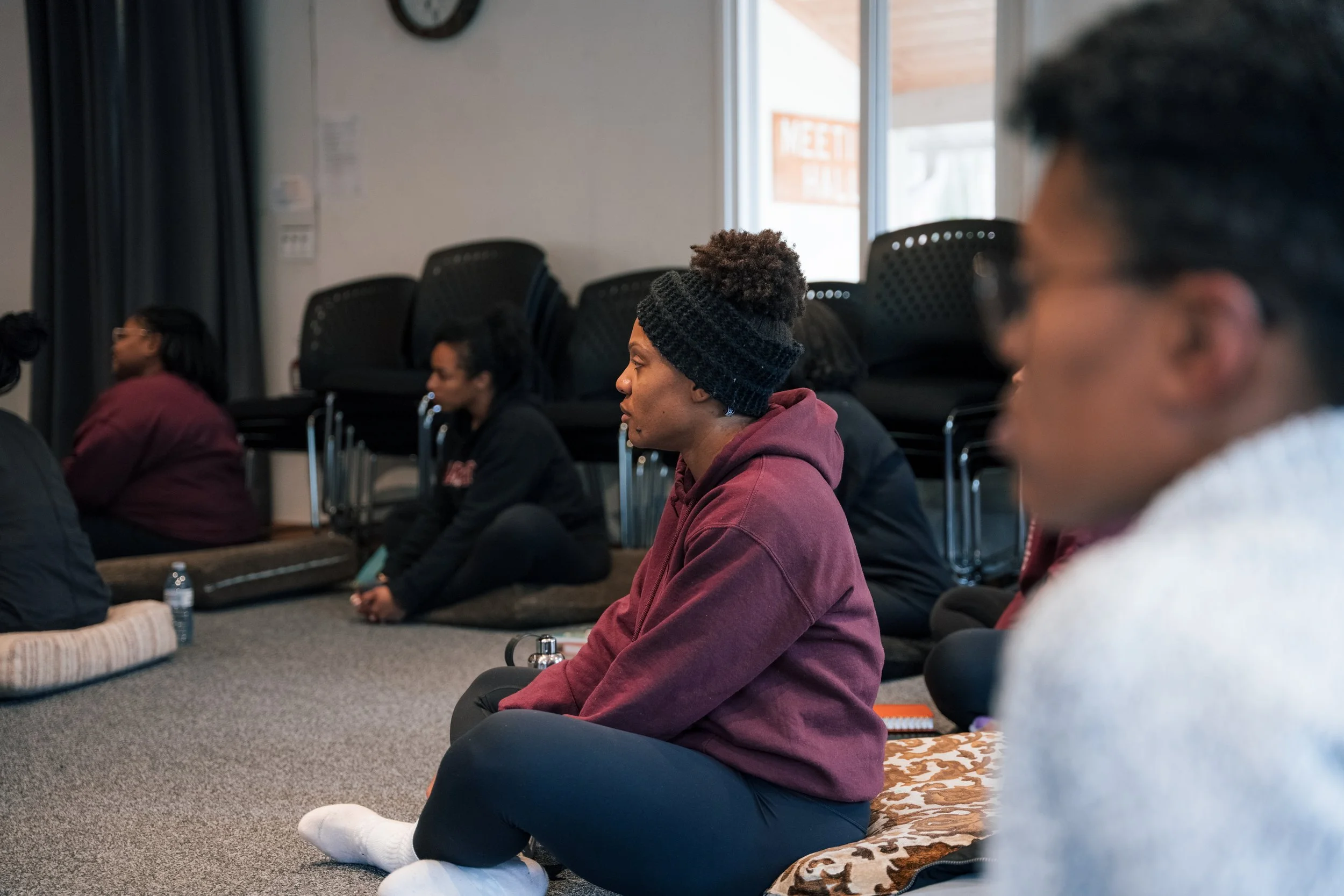 Group of people sitting cross-legged on the floor during a meditation or mindfulness session in a room with black chairs stacked against the wall and a window.