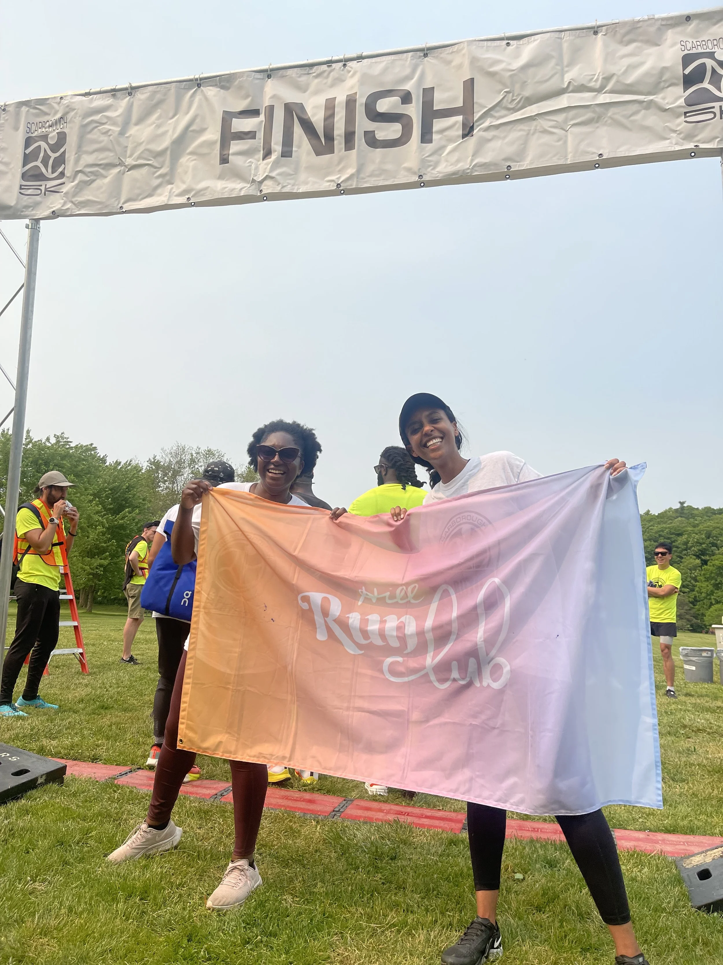 Two women smiling and holding a large pink and white flag that reads 'Hill Run Club' in front of a finish line banner at an outdoor event, with several people in bright clothing and green trees in the background.
