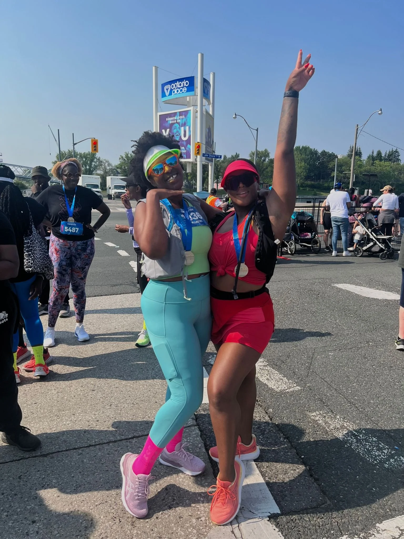 Two women celebrating after a race, wearing running gear and medals, standing on a city street with other runners and spectators in the background.