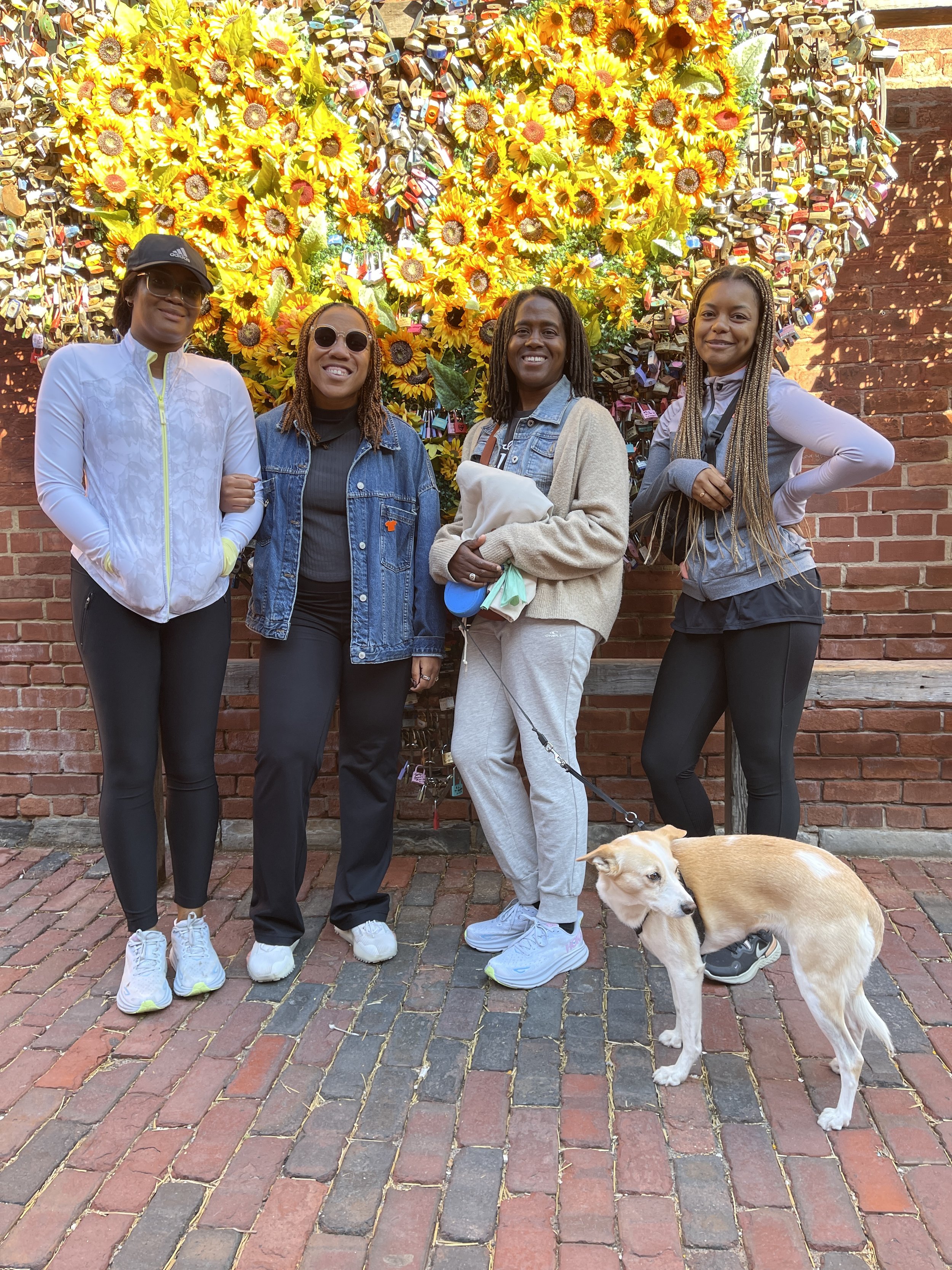 Four women and a dog standing in front of a wall decorated with yellow sunflowers and numerous padlocks, on a brick-paved street during daytime.
