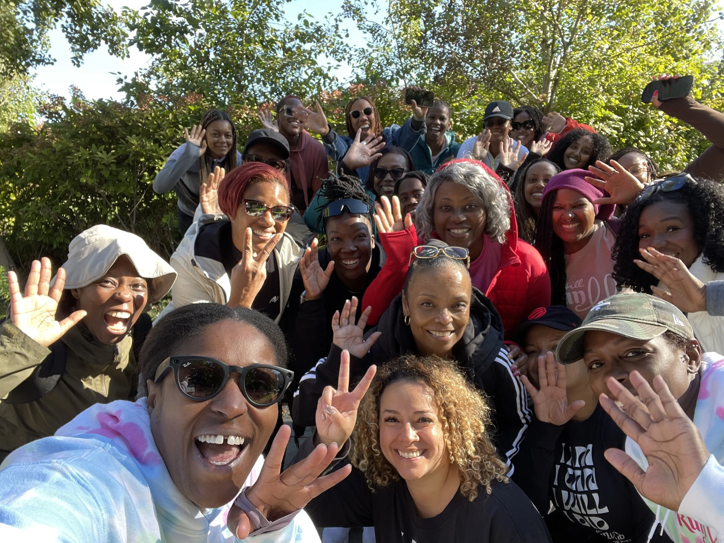 Large group of diverse people smiling and waving outdoors during daytime, some holding phones, gathered in front of green trees.