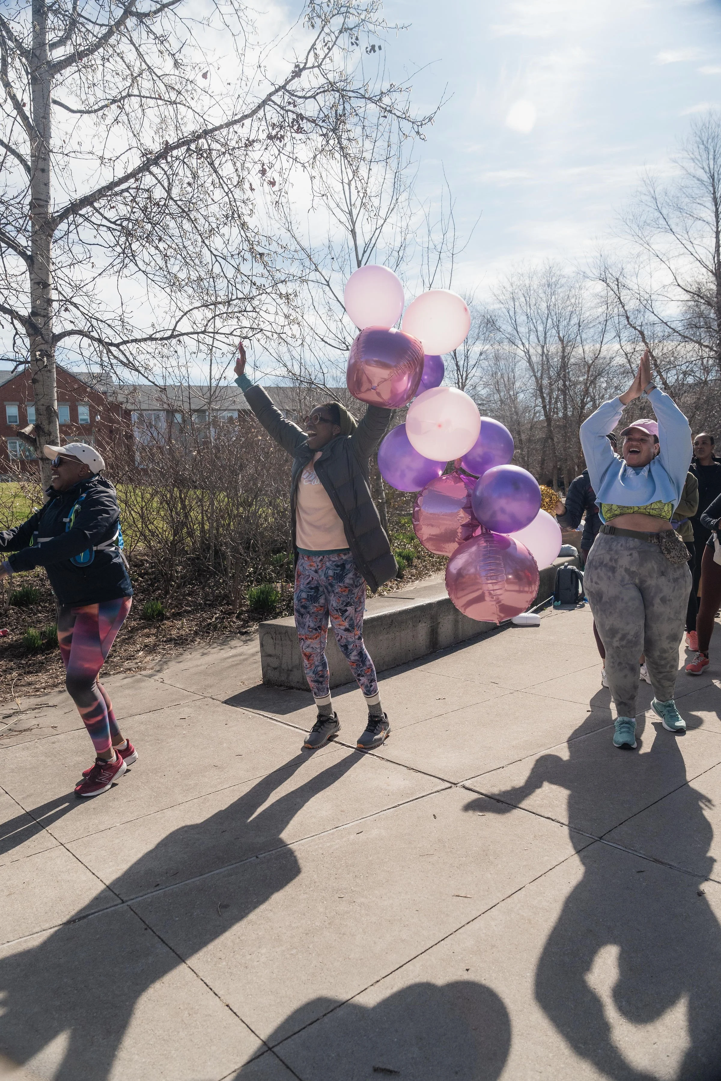 Group of people dancing outdoors on a sunny day, with some holding balloons, among leafless trees and residential buildings in the background.