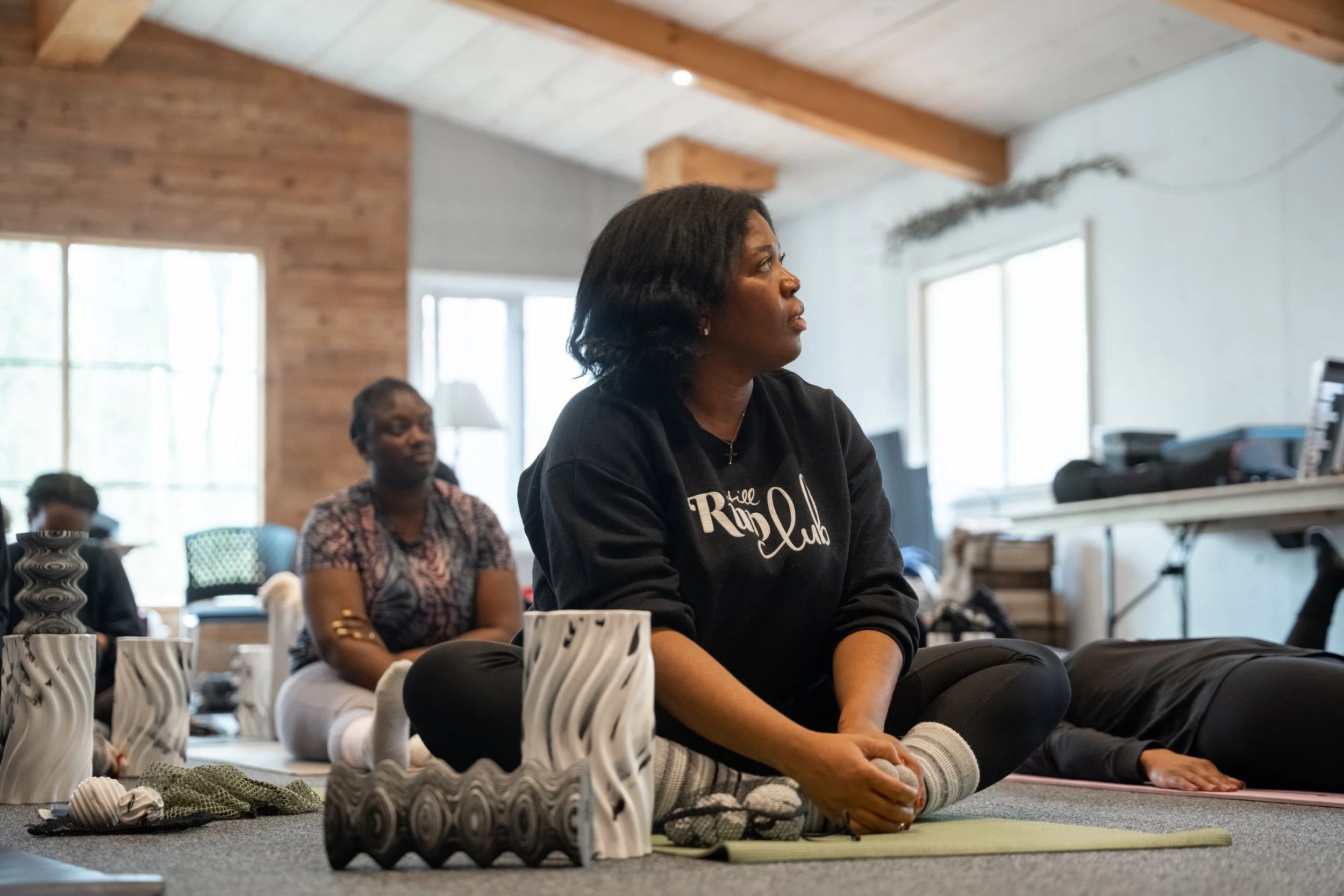 Women participating in a yoga or meditation class in a room with large windows and wooden ceiling beams, sitting cross-legged on mats.