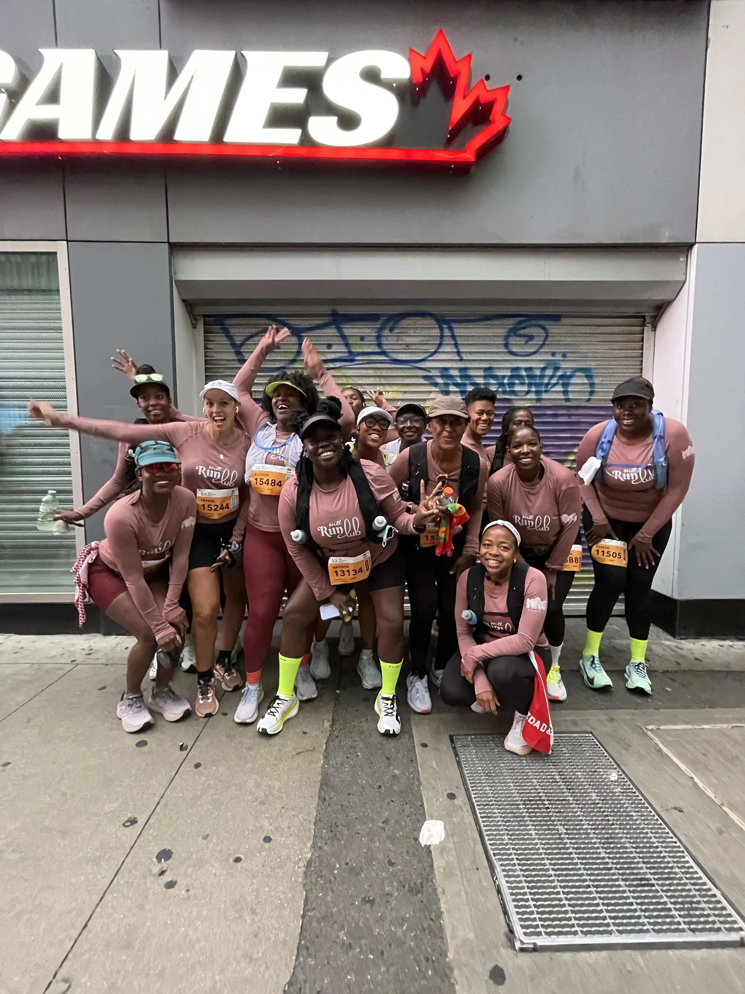 Group of runners in pink shirts posing in front of a closed storefront with graffiti, celebrating after a race.