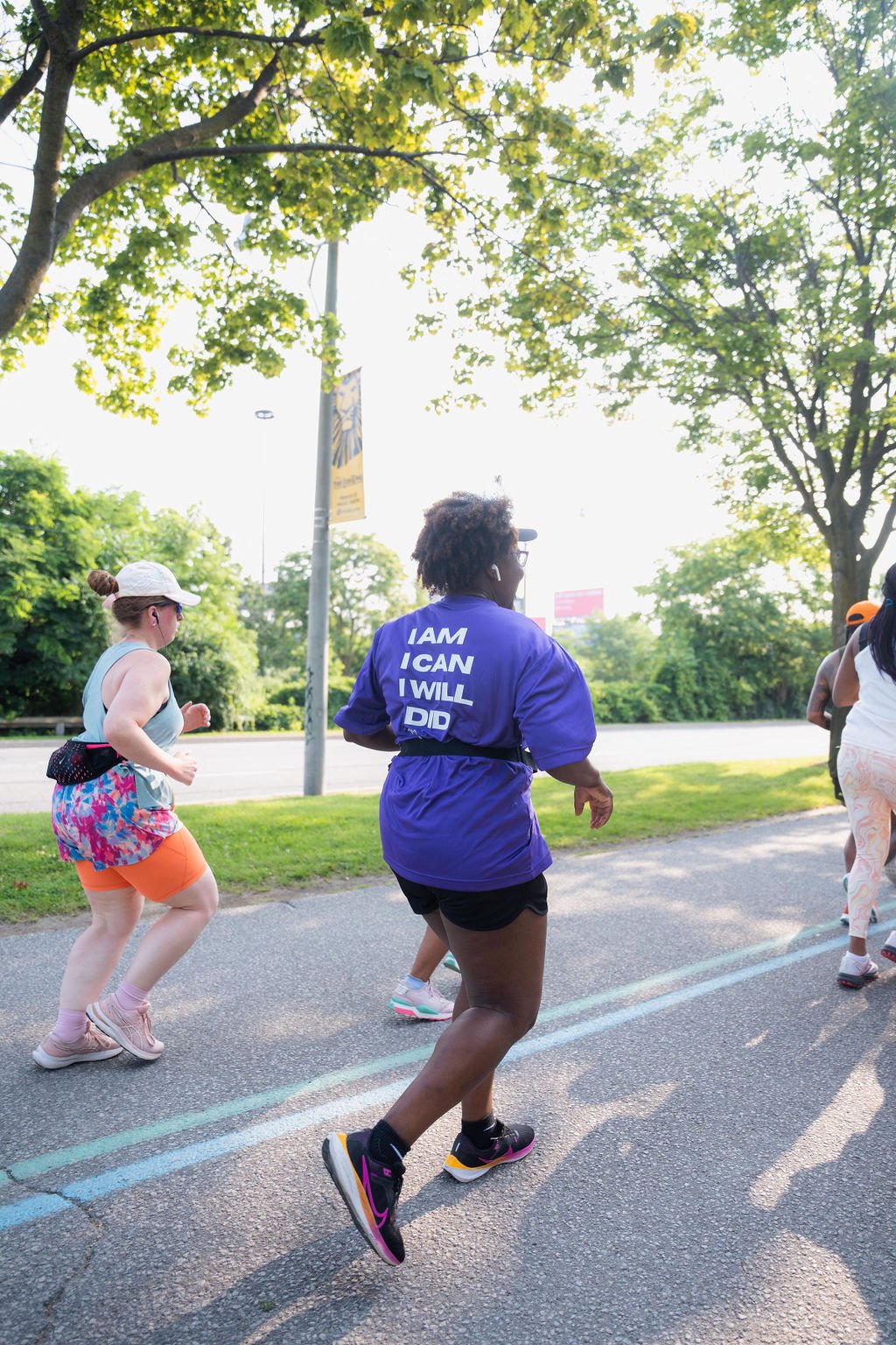 People jogging on a tree-lined street during daytime, with a woman in a purple shirt that reads 'I AM I CAN I WILL DID'.