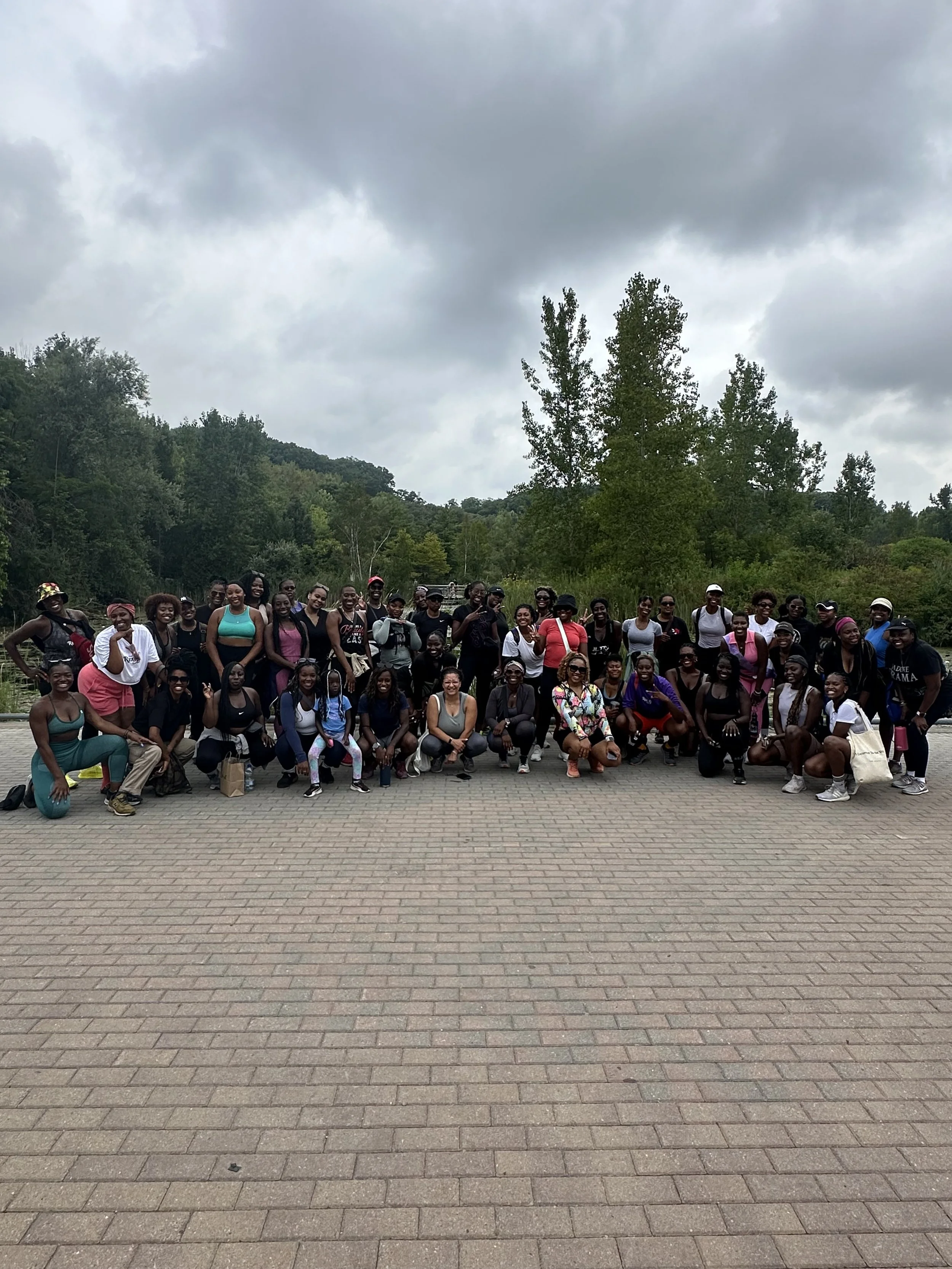 A large group of women posing outdoors on a paved area with trees and hills in the background under cloudy skies.