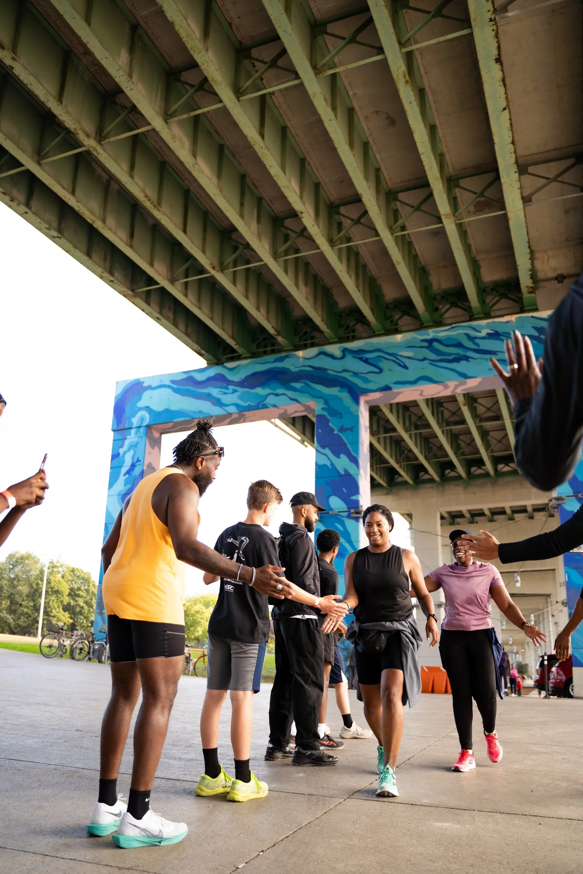 People at an outdoor event under a bridge, with some giving high-fives and others taking photos, in front of a colorful blue and pink painted frame.