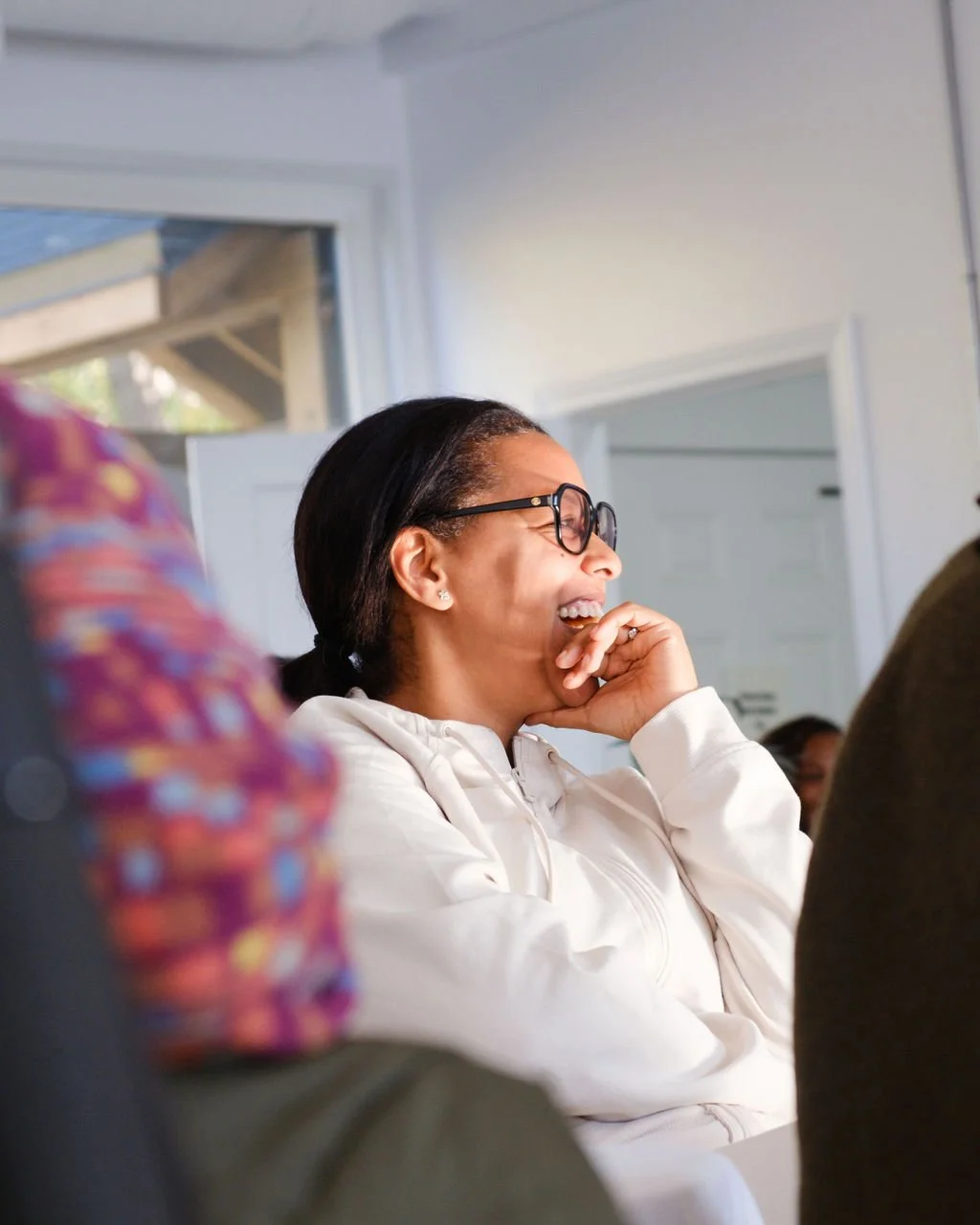 A woman with glasses and earrings laughing and covering her mouth with her hand during a gathering or meeting.