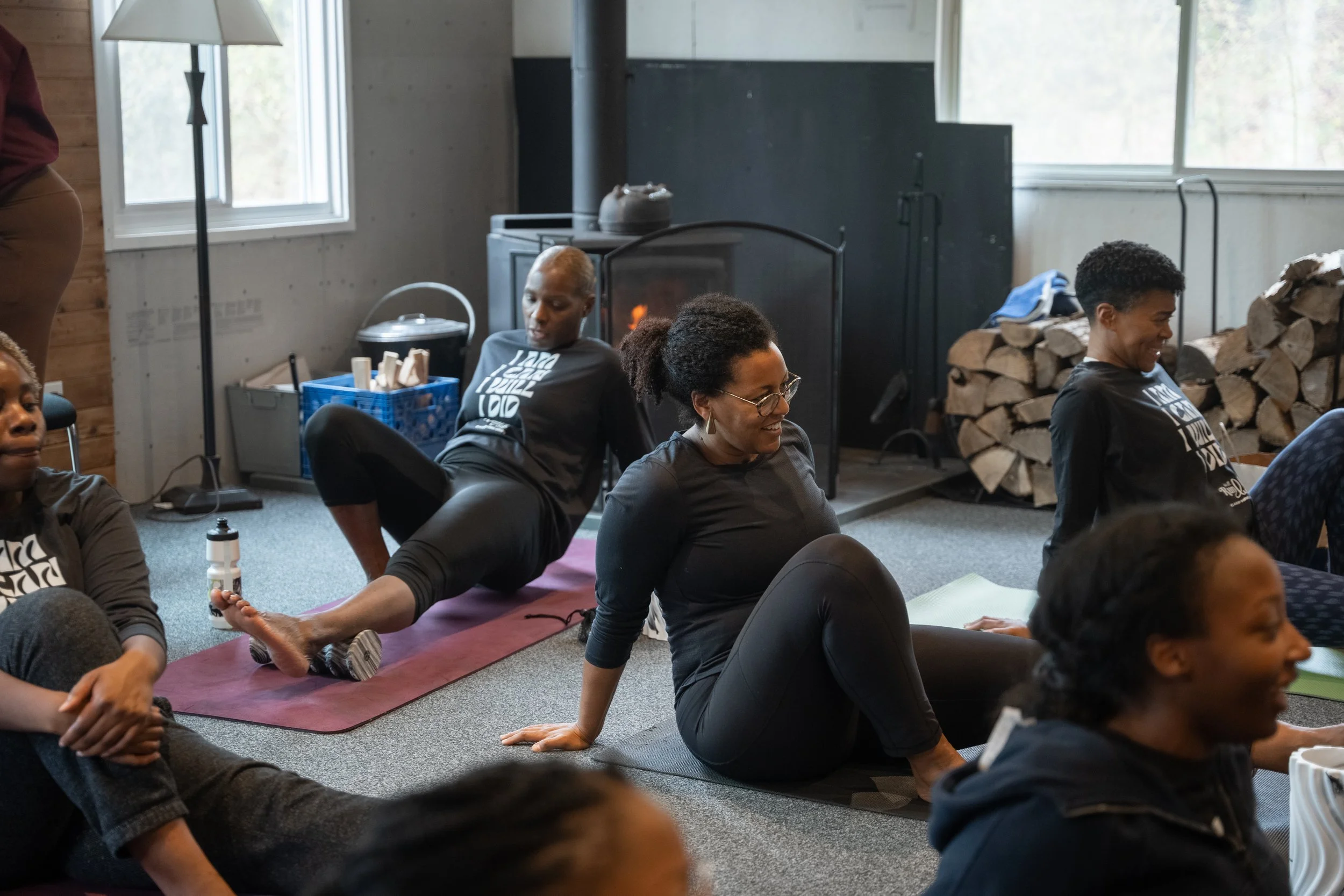 Group of people practicing yoga indoors, sitting on mats, with a wood-burning stove and logs in the background.