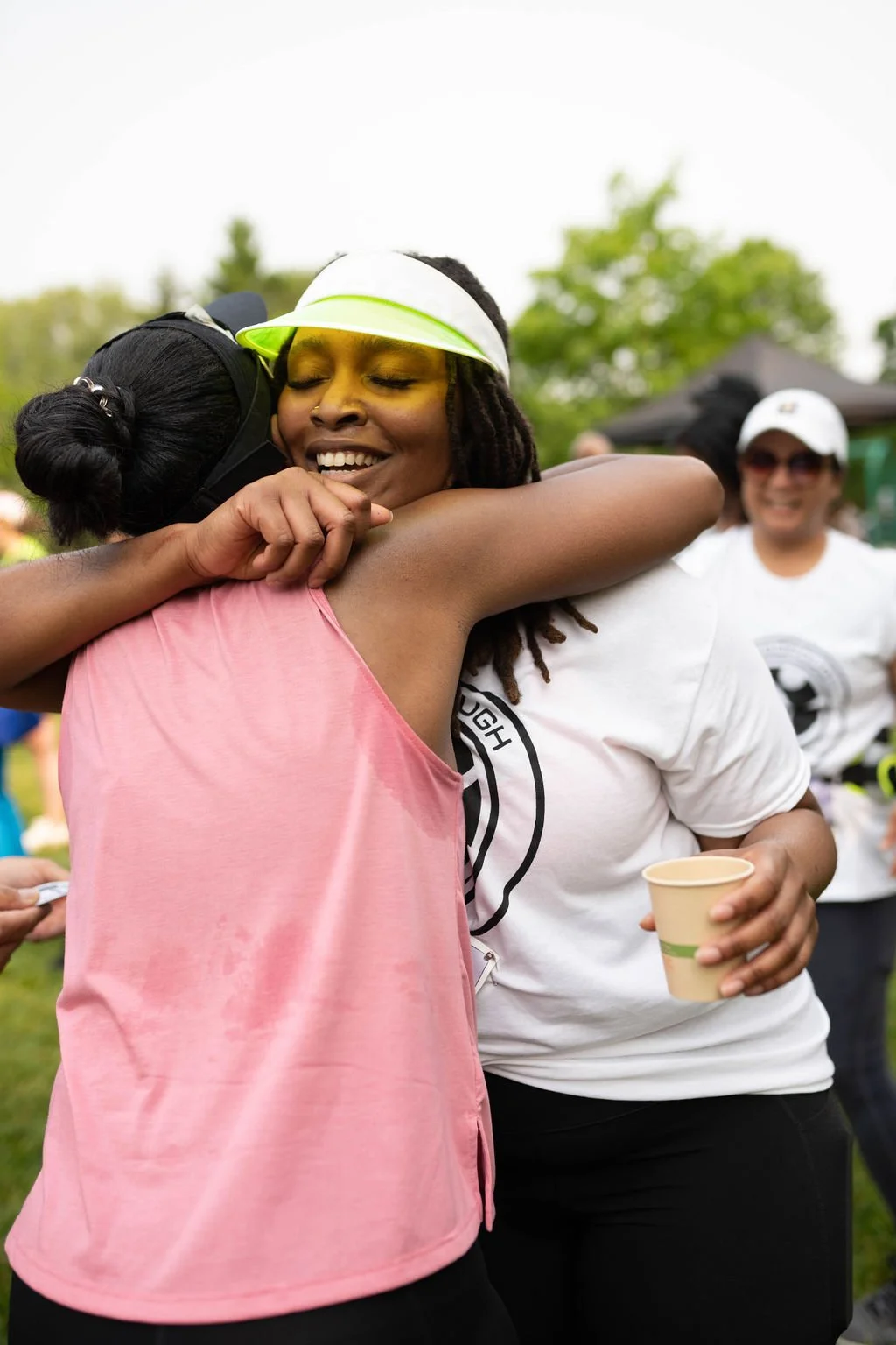 Two women hugging each other at a park event, one wearing a pink tank top and the other wearing a white t-shirt with a logo, holding a paper cup, smiling and joyful, with other people in the background.