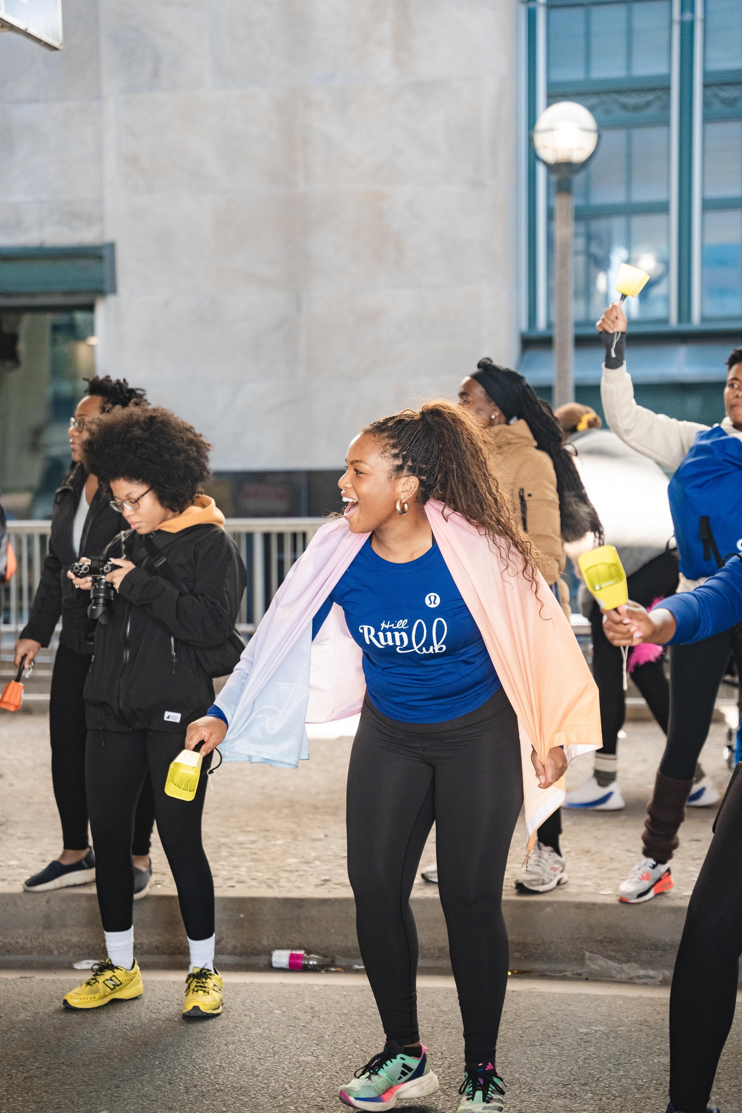 Woman wearing a blue shirt with 'Still Run Club' text, laughing, surrounded by people with small yellow batons, at an outdoor event, possibly a run or walk event, on a city street.