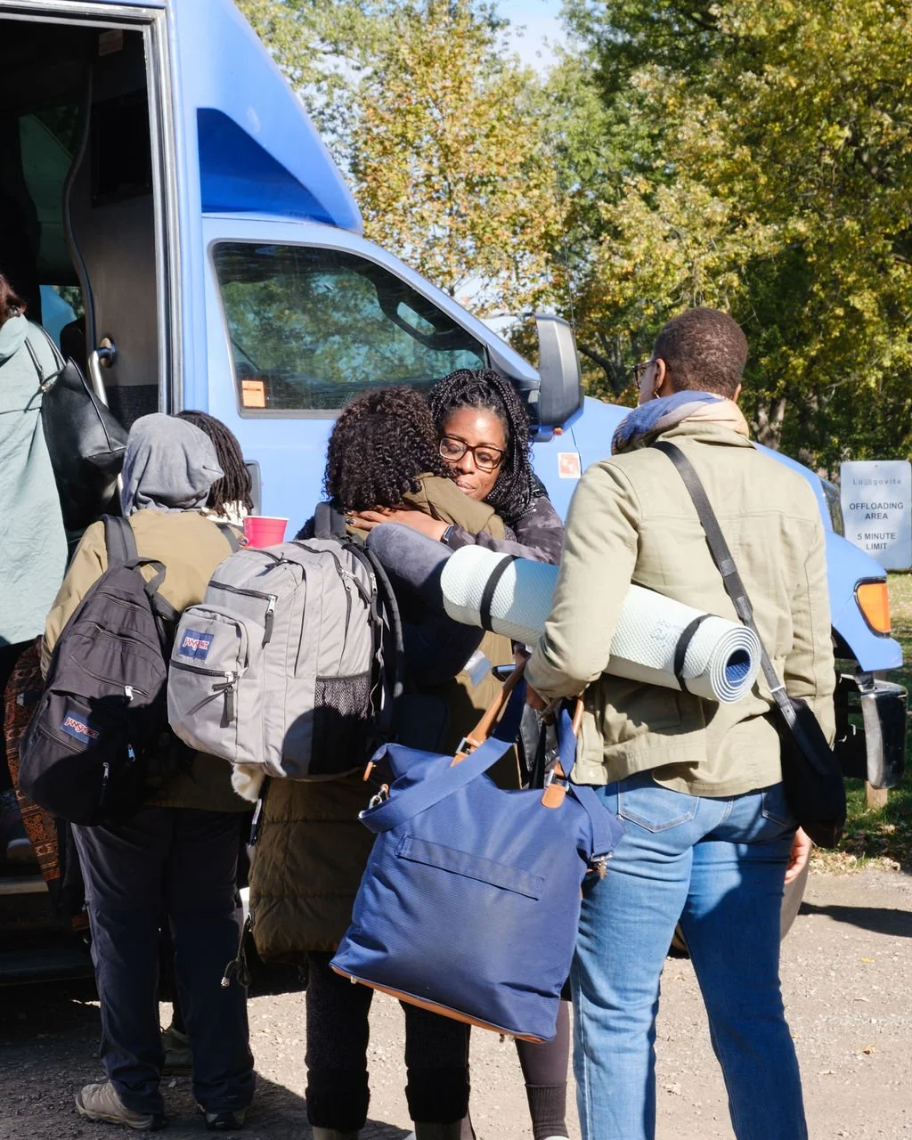 People embracing in front of a blue truck outdoors on a sunny day, with trees in the background.