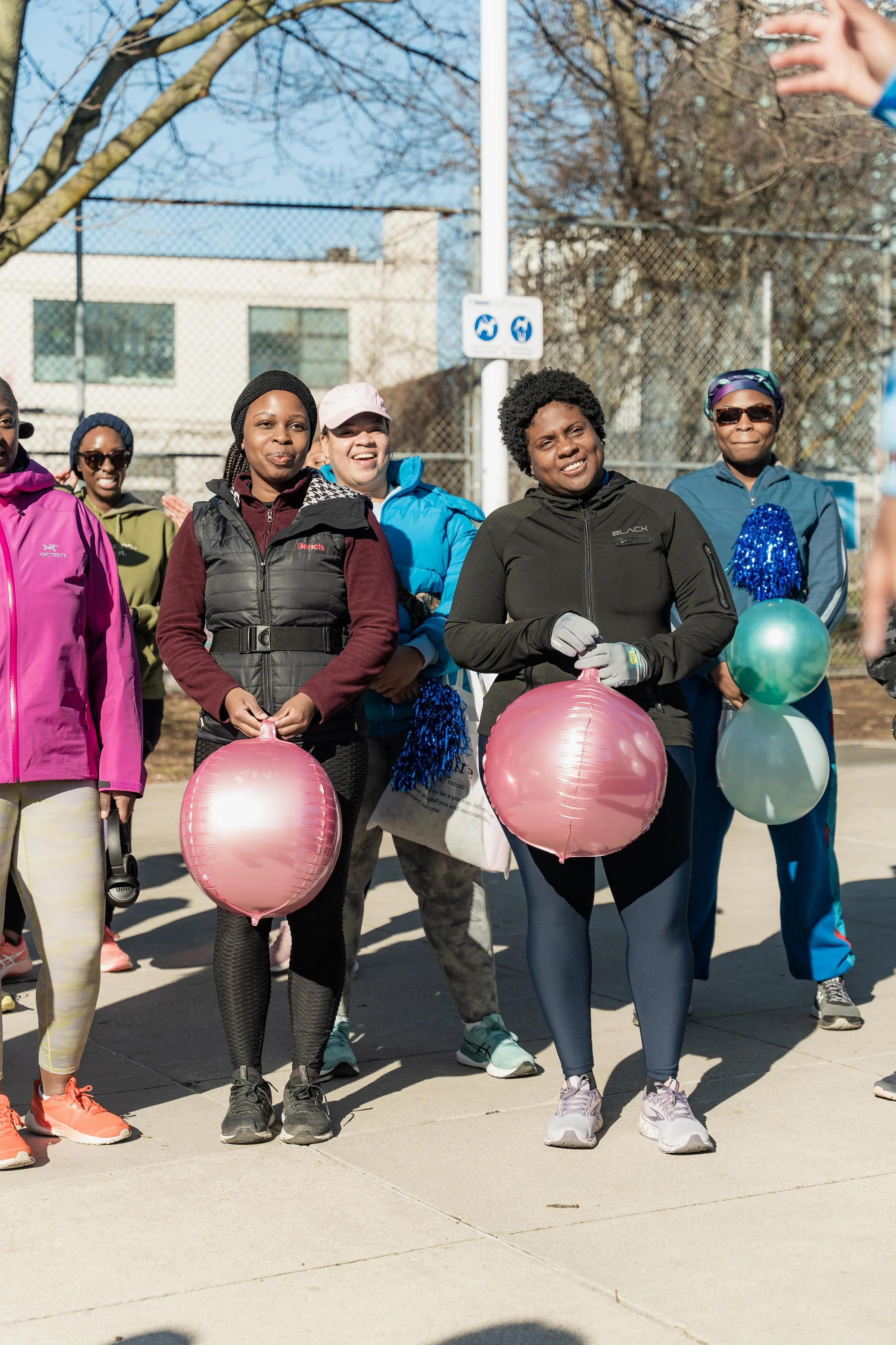 Group of women standing outdoors, holding pink and green balloons, dressed in athletic clothing, on a sunny day at a park or sports court.