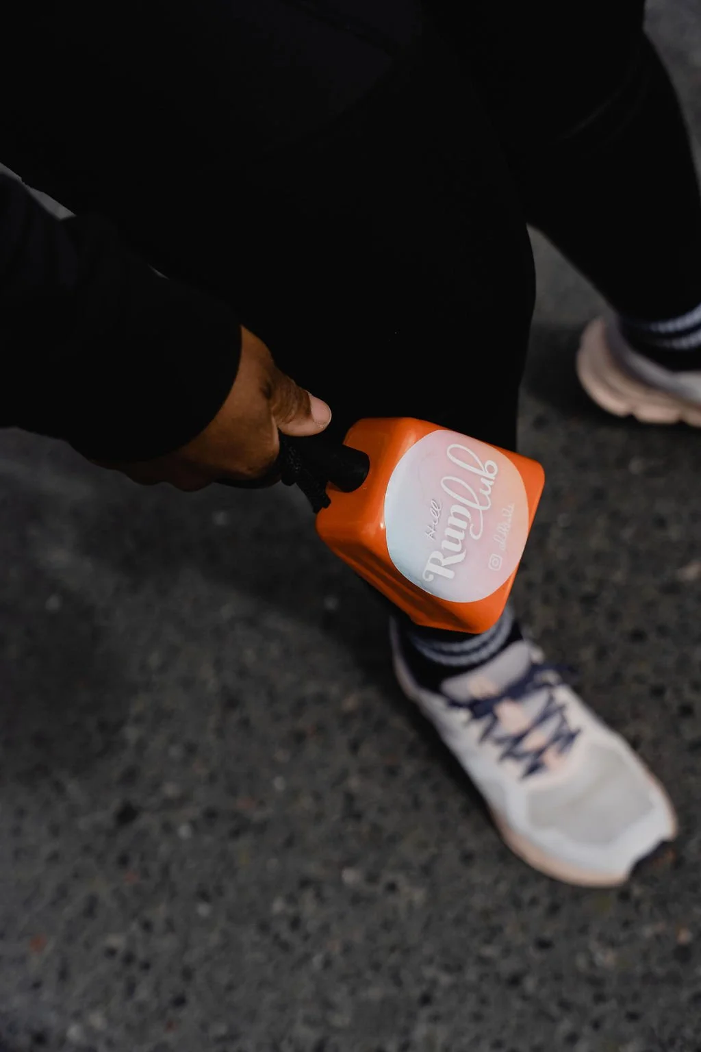 Person holding an orange water bottle with white label that reads 'Run Club' and a sneaker on a paved ground.