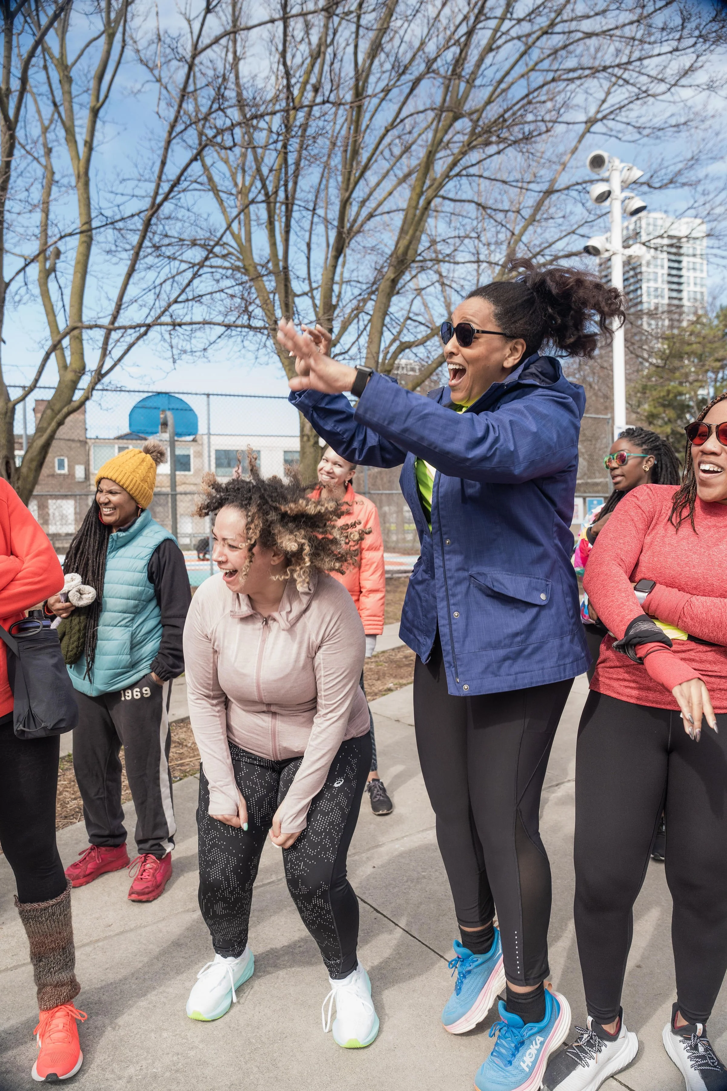Group of diverse women laughing and having fun during outdoor event on a sunny day, with sports court, trees, and city buildings in background.