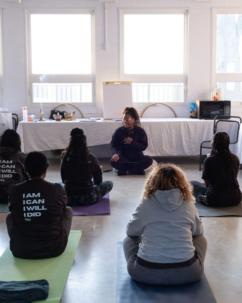 Group of women participating in a yoga or meditation class in a bright room with large windows, seated on mats facing an instructor sitting cross-legged on the floor.