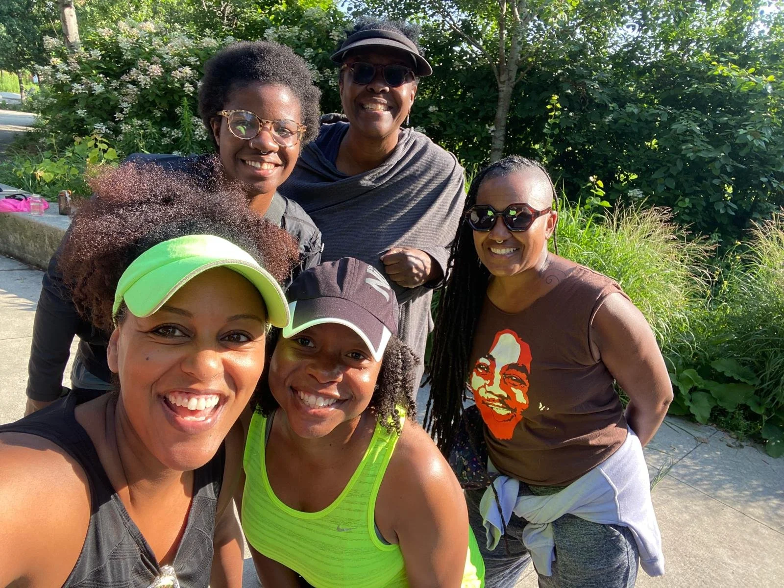 Group of six women smiling outdoors, wearing athletic clothing and sunglasses, on a sunny day with green trees and bushes in the background.