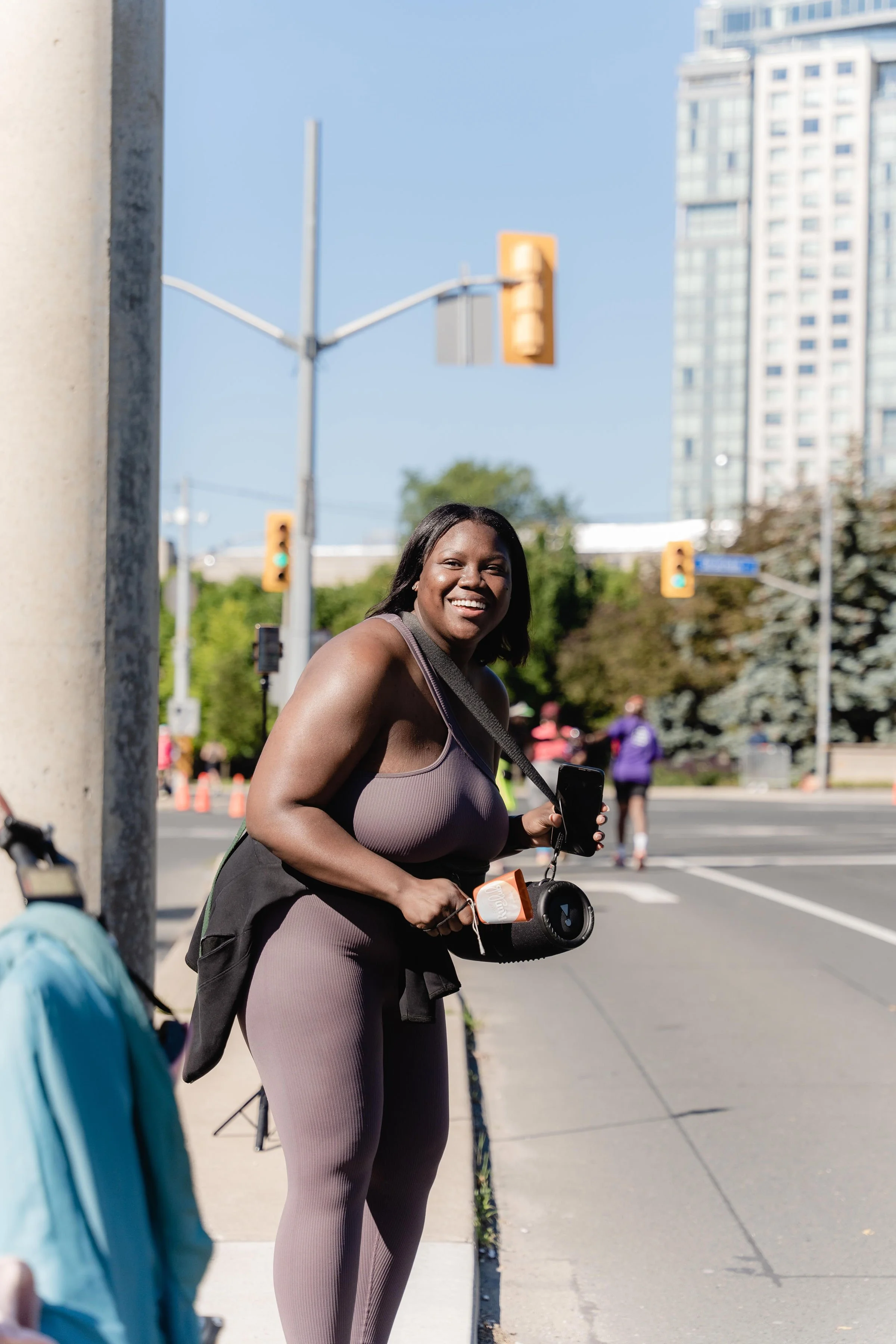 A woman smiling on a city sidewalk, holding a phone and a speaker, with people and traffic lights in the background.