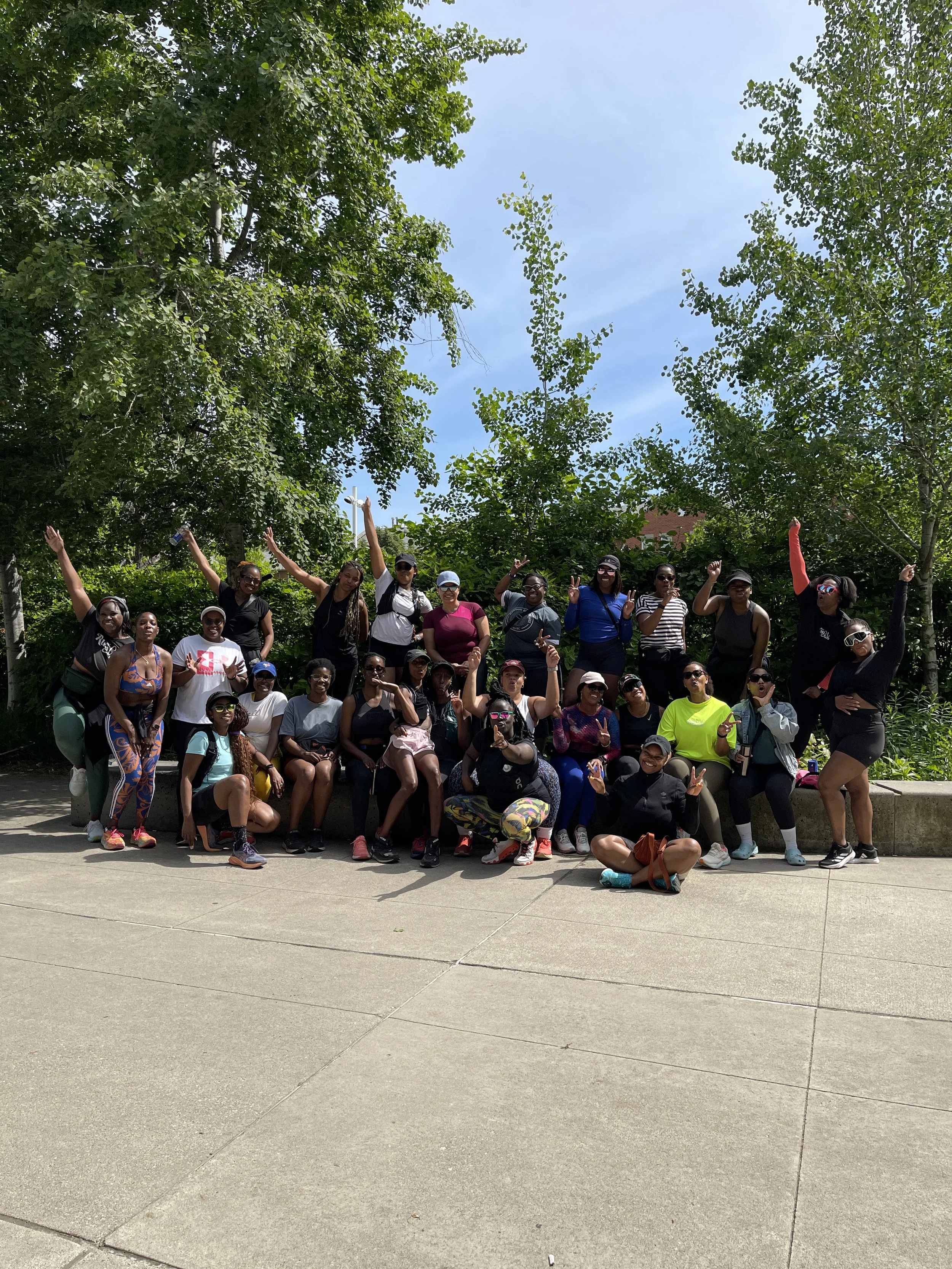Group of women and girls posing outdoors on a sunny day, some making peace signs, in front of greenery and trees.