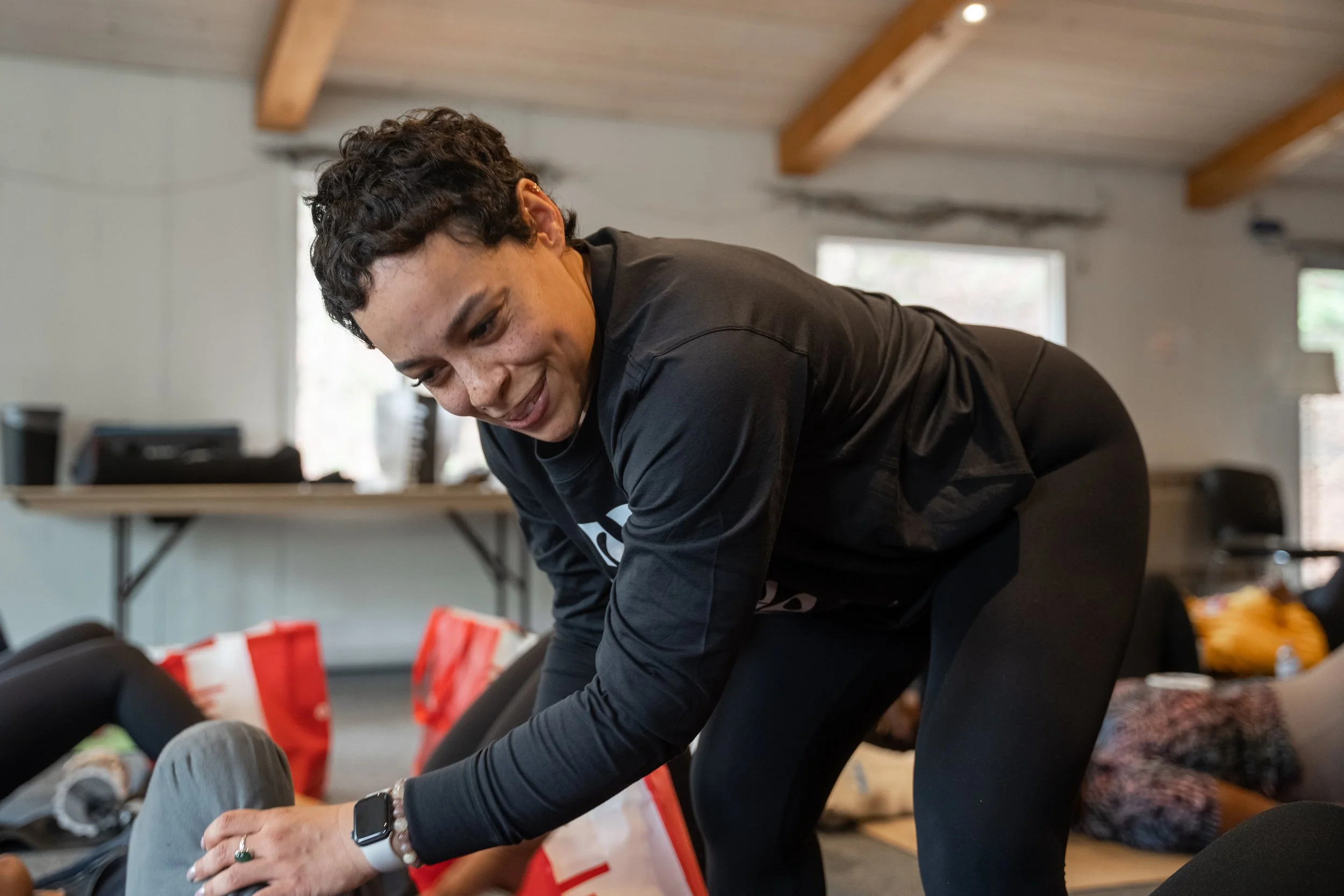 A woman with curly dark hair, wearing a black long-sleeved shirt and black leggings, is smiling and leaning forward as she helps another person during an indoor activity.