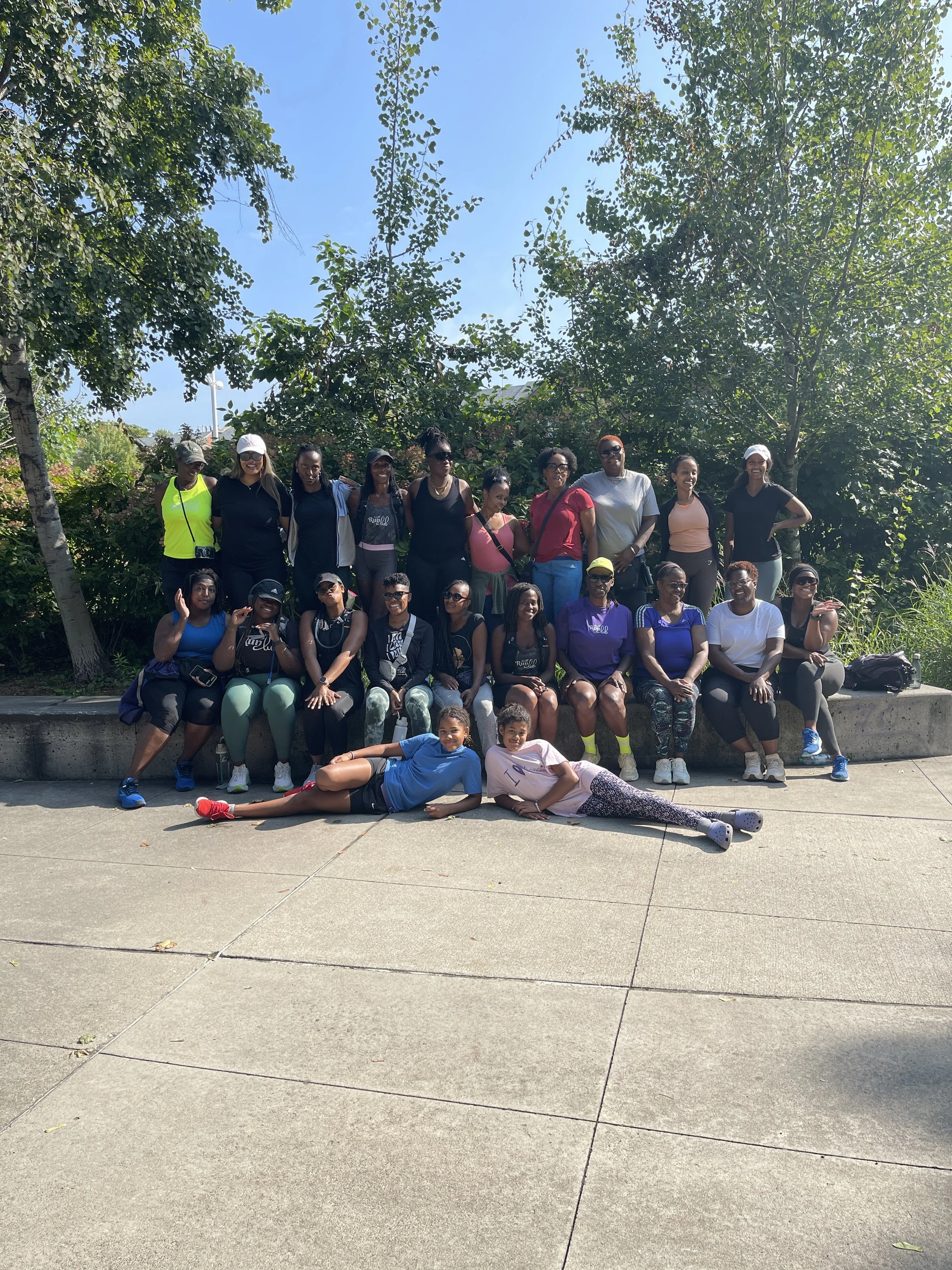 Group of diverse women and children outdoors on a sunny day, sitting and standing in front of trees.
