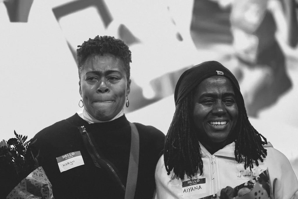 Two women standing together, one crying and the other smiling, both wearing name tags, in an indoor setting.