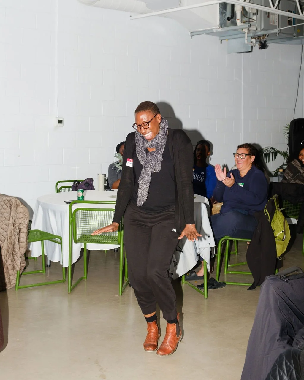 A woman wearing glasses, a black outfit, and brown boots is dancing joyfully with a big smile, while a group of women seated at tables in the background cheer and clap in a well-lit indoor setting.