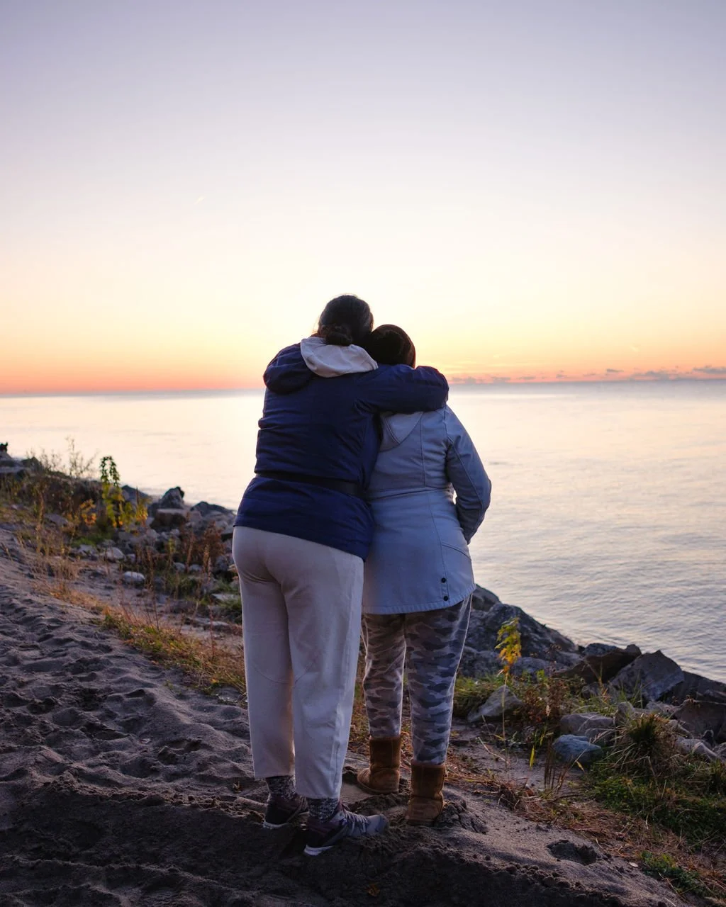 Two people embrace while watching a sunset over the ocean from a rocky beach.
