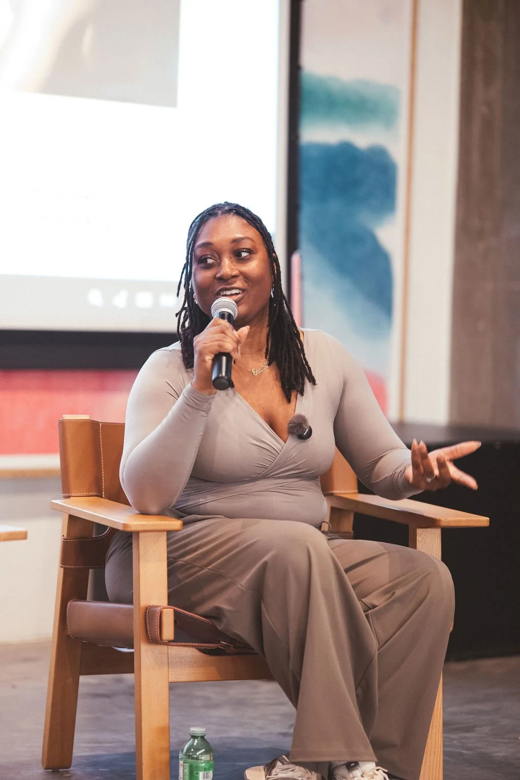 A woman with dreadlocks speaking into a microphone during a panel discussion or interview, seated on a wooden chair with a water bottle on the floor nearby.