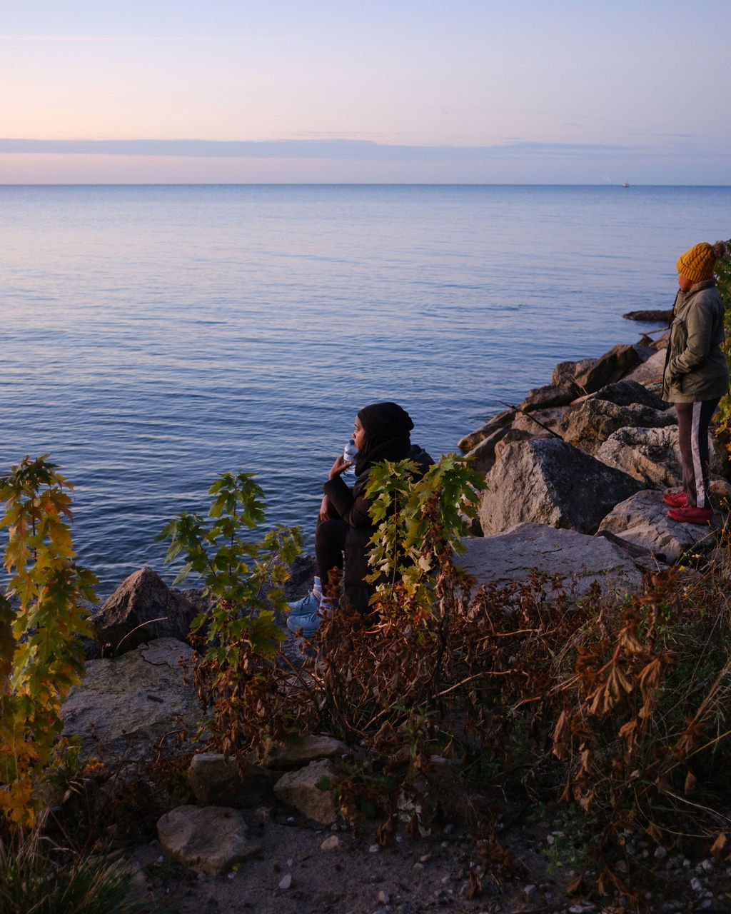 Two children on rocky shoreline by a calm body of water during sunset, one sitting and drinking water, the other standing with fishing rod, surrounded by foliage.