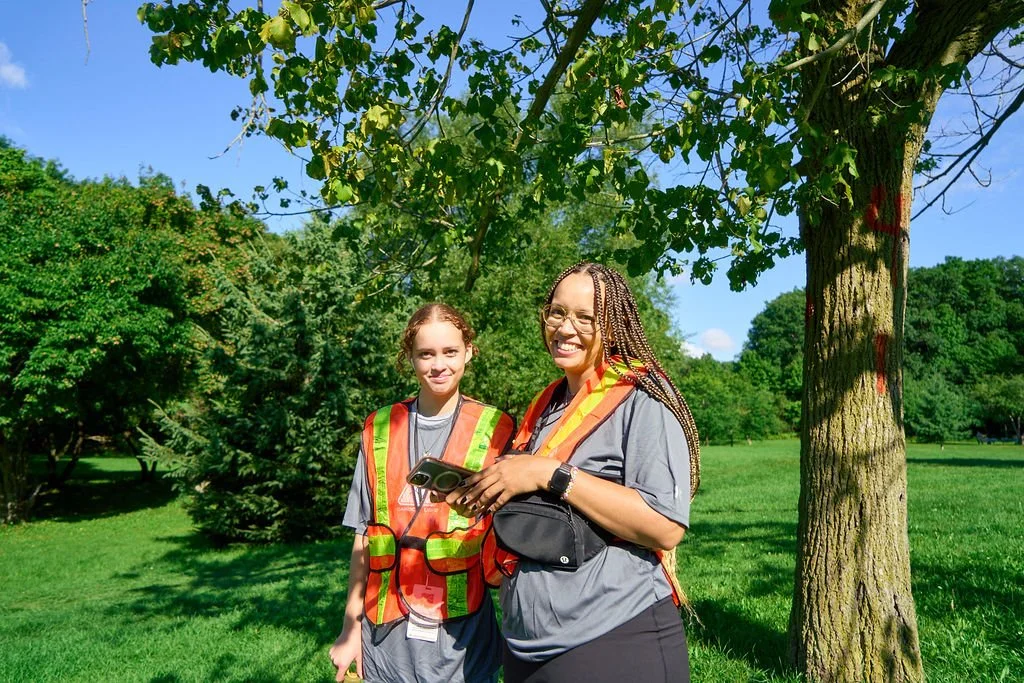 Two women standing outdoors under a tree, smiling at the camera. They are wearing gray shirts and safety vests, with one holding a phone.