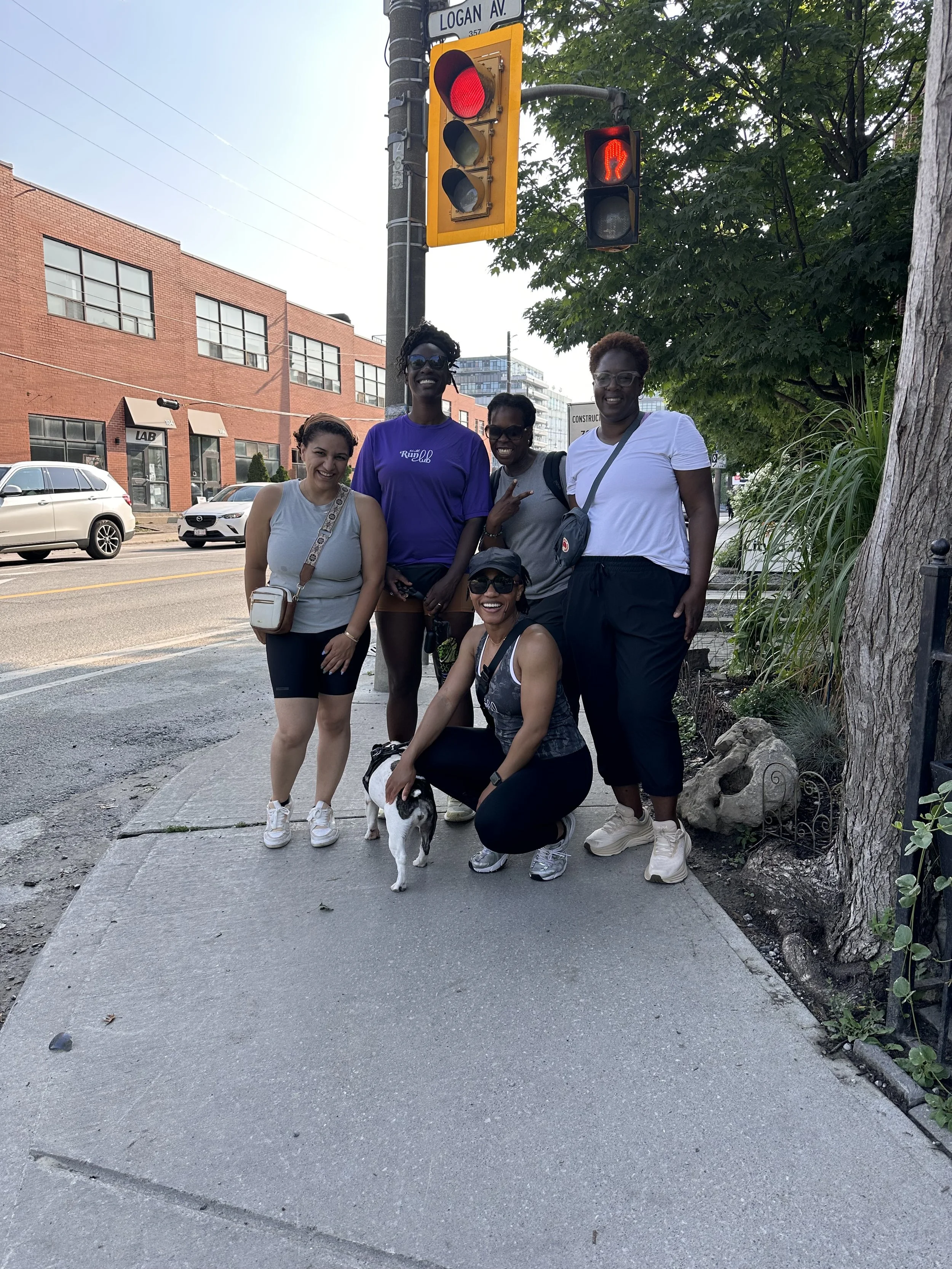 Group of five women posing at a street corner with a small dog, traffic light showing red pedestrian signal, and trees in the background.