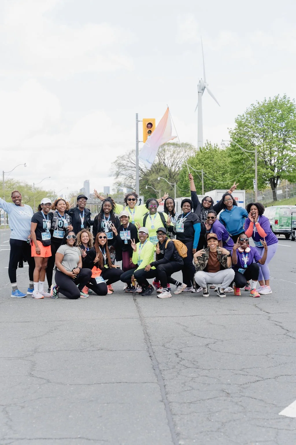 Group of diverse volunteers or participants at a race or outdoor event, posing cheerfully, some holding flags and making peace signs, with a wind turbine and city skyline in the background.