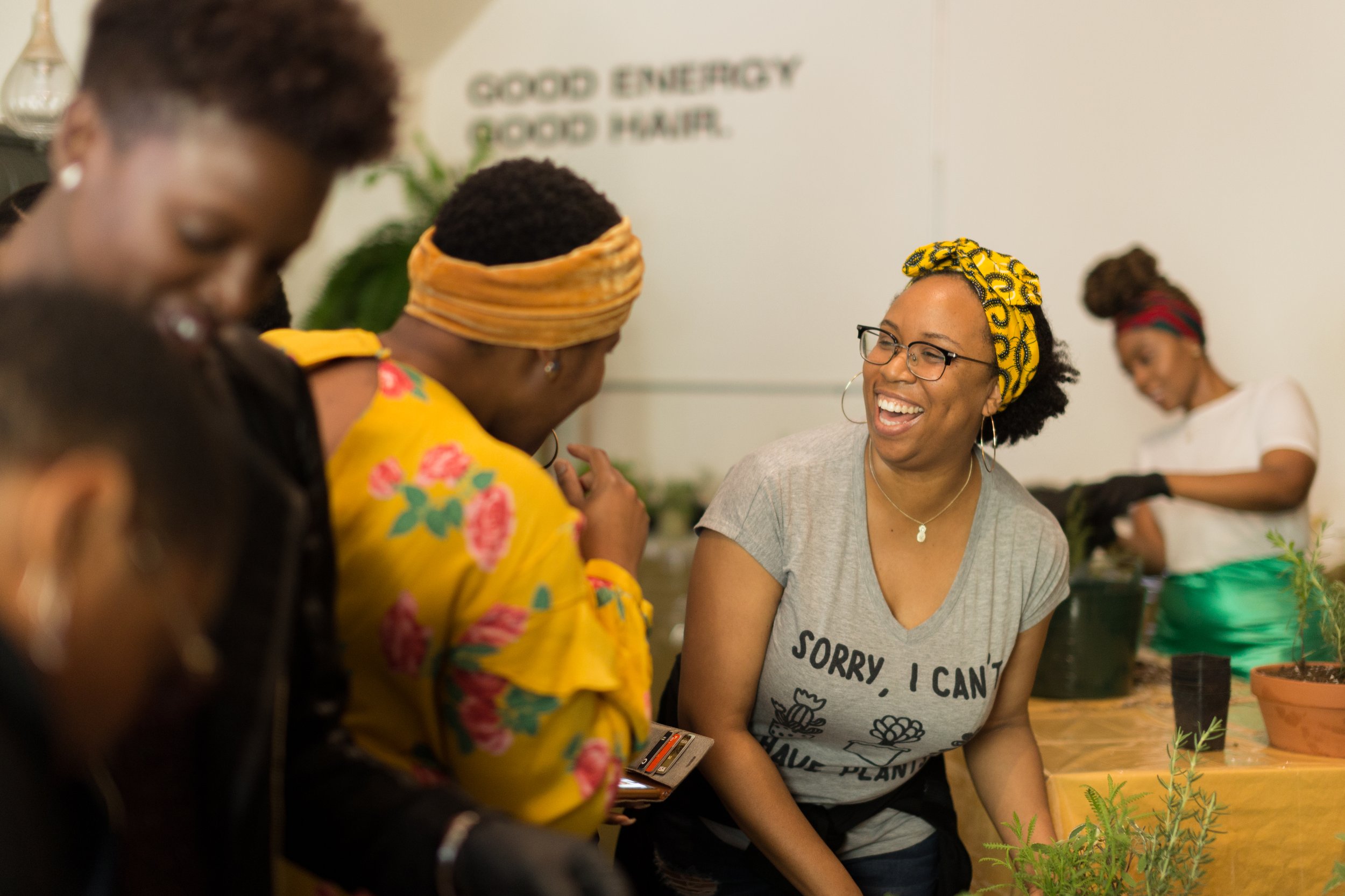 A group of women engaging in a gardening activity indoors, some are wearing gloves and aprons, with smiling and laughing. One woman stands out with a yellow headscarf and glasses, wearing a gray t-shirt that says 'Sorry, I can't, I have plans' and is