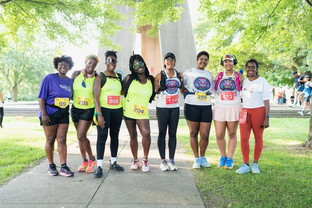 Group of women in athletic gear, standing outdoors on a sunny day, after a race or marathon, smiling and posing for the photo.