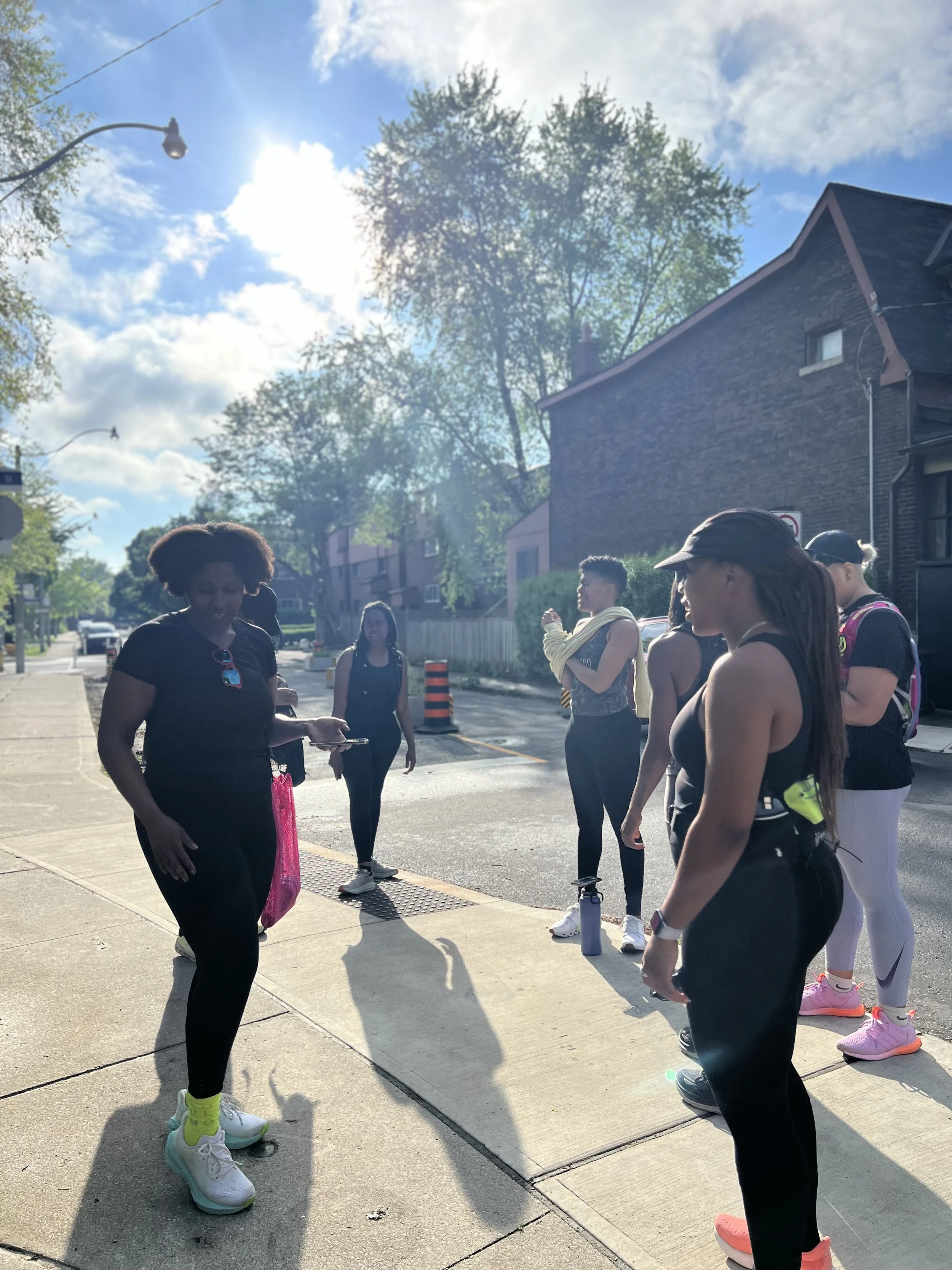 A group of women in athletic clothing gathered on a sidewalk in a residential neighborhood, preparing for a walk or run on a sunny day.