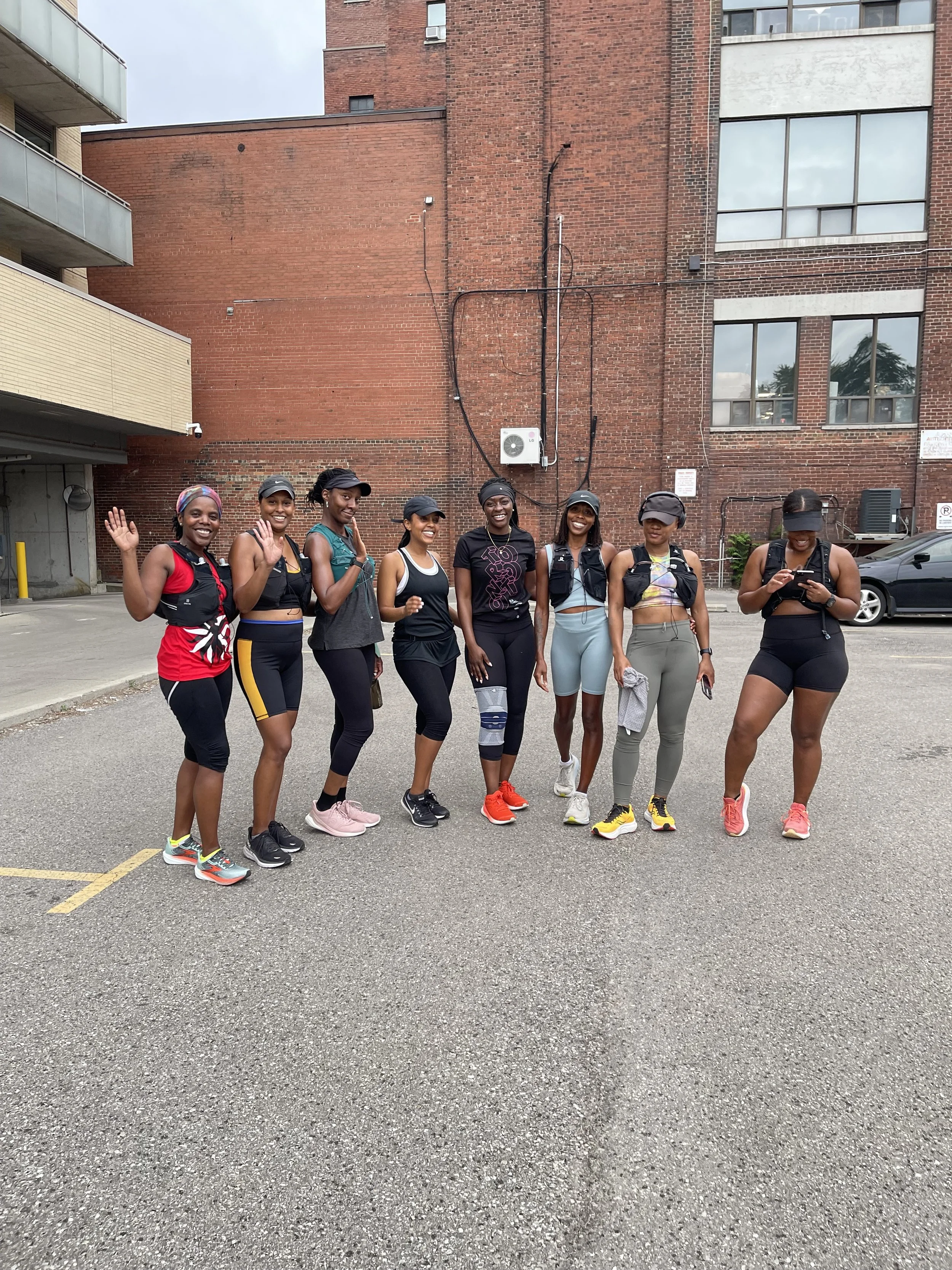 Eight women in athletic clothing standing together outdoors in a parking lot, smiling and waving at the camera with a brick building in the background.