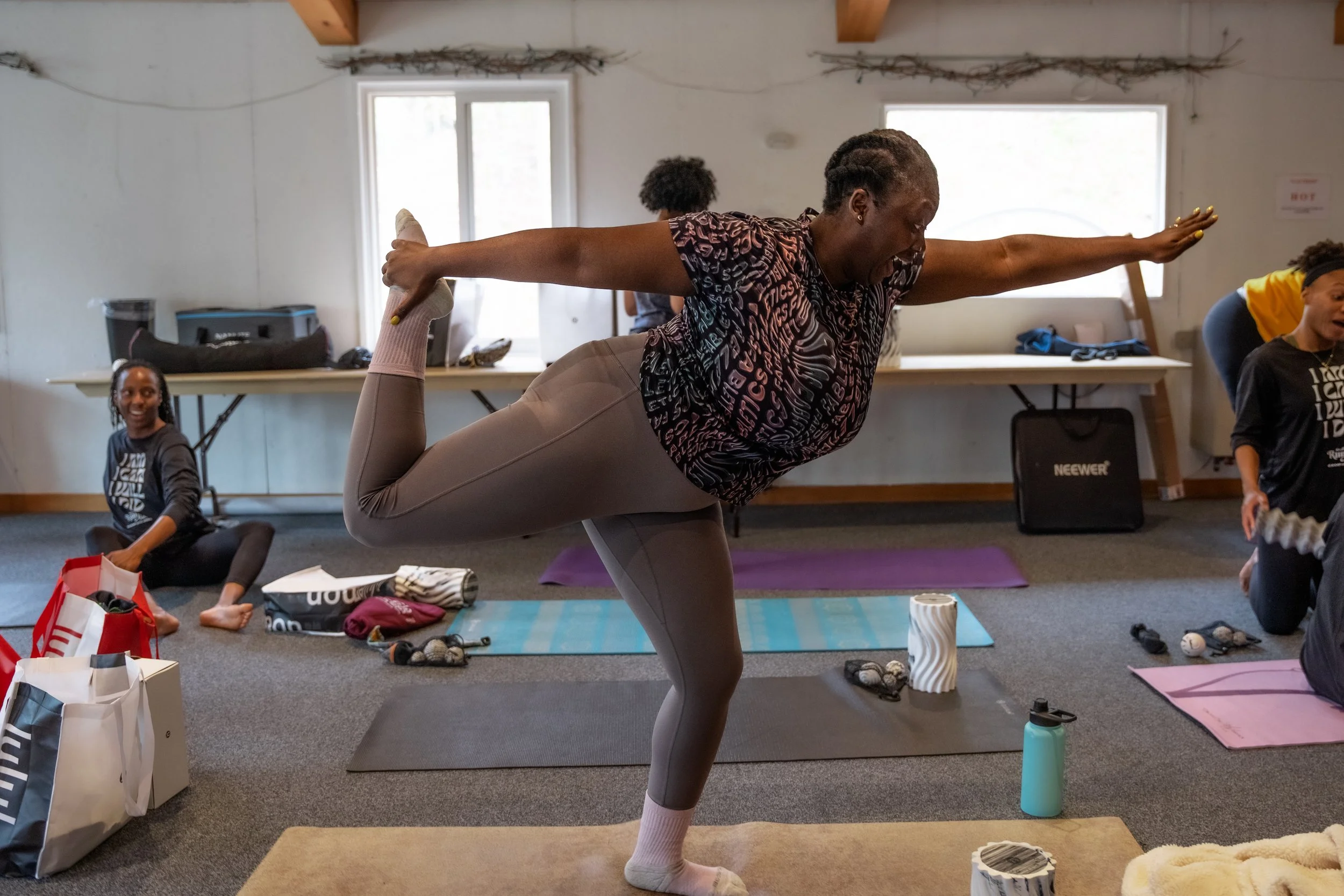 Woman practicing yoga in a class with other participants following her pose, indoor setting with yoga mats and supplies.
