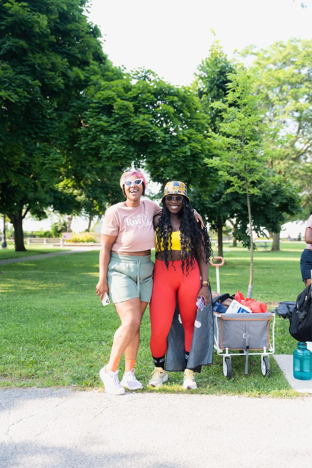 Two smiling women standing outdoors in a park, with trees and greenery in the background, one wearing a pink shirt and shorts, the other in a yellow top and red pants, holding a wagon filled with items.
